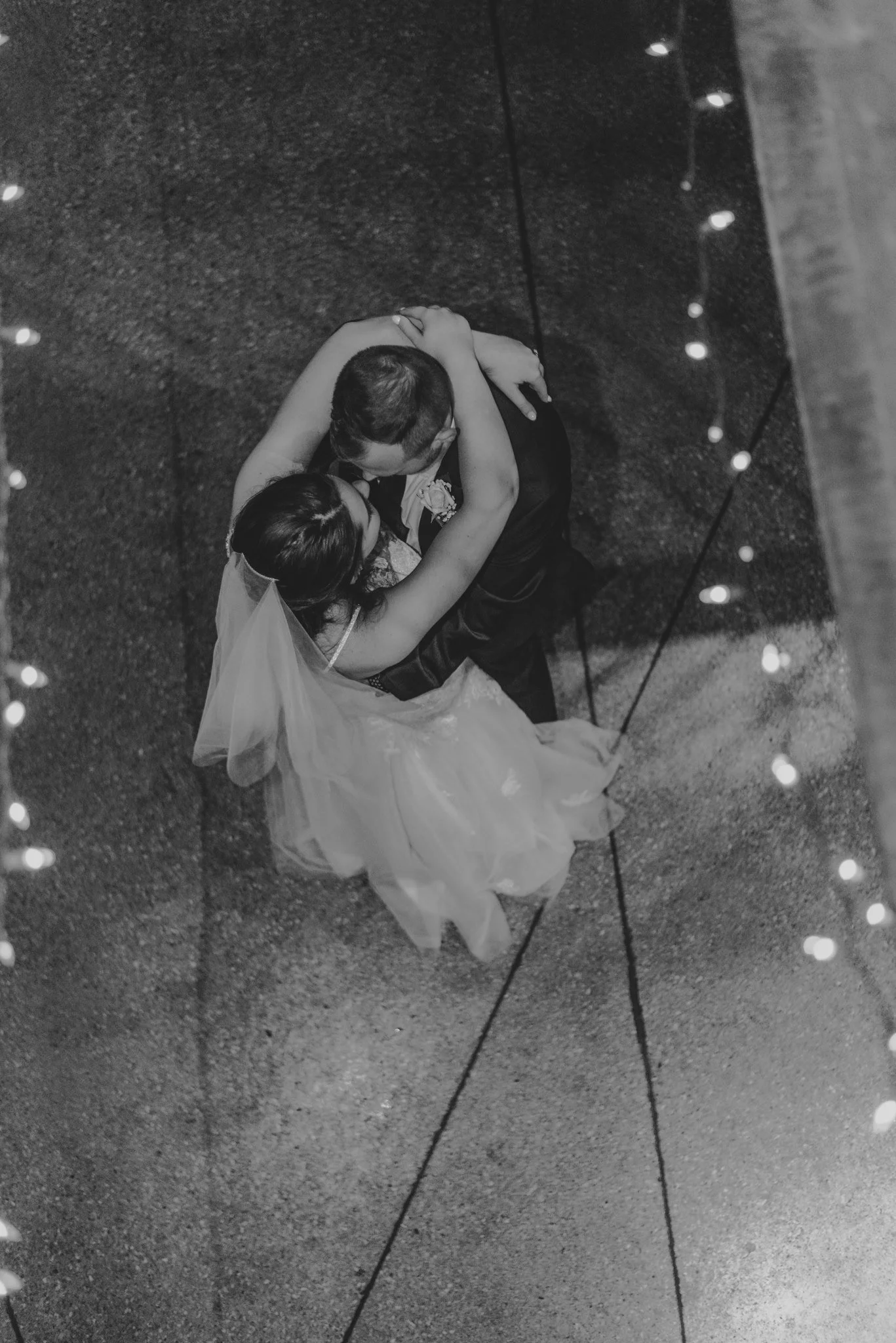 A black-and-white photo of a newlywed couple dancing their first dance, captured from above. The bride is wearing a wedding dress and veil, and the groom is in a dark suit. They are embracing each other on a textured floor with string lights around t