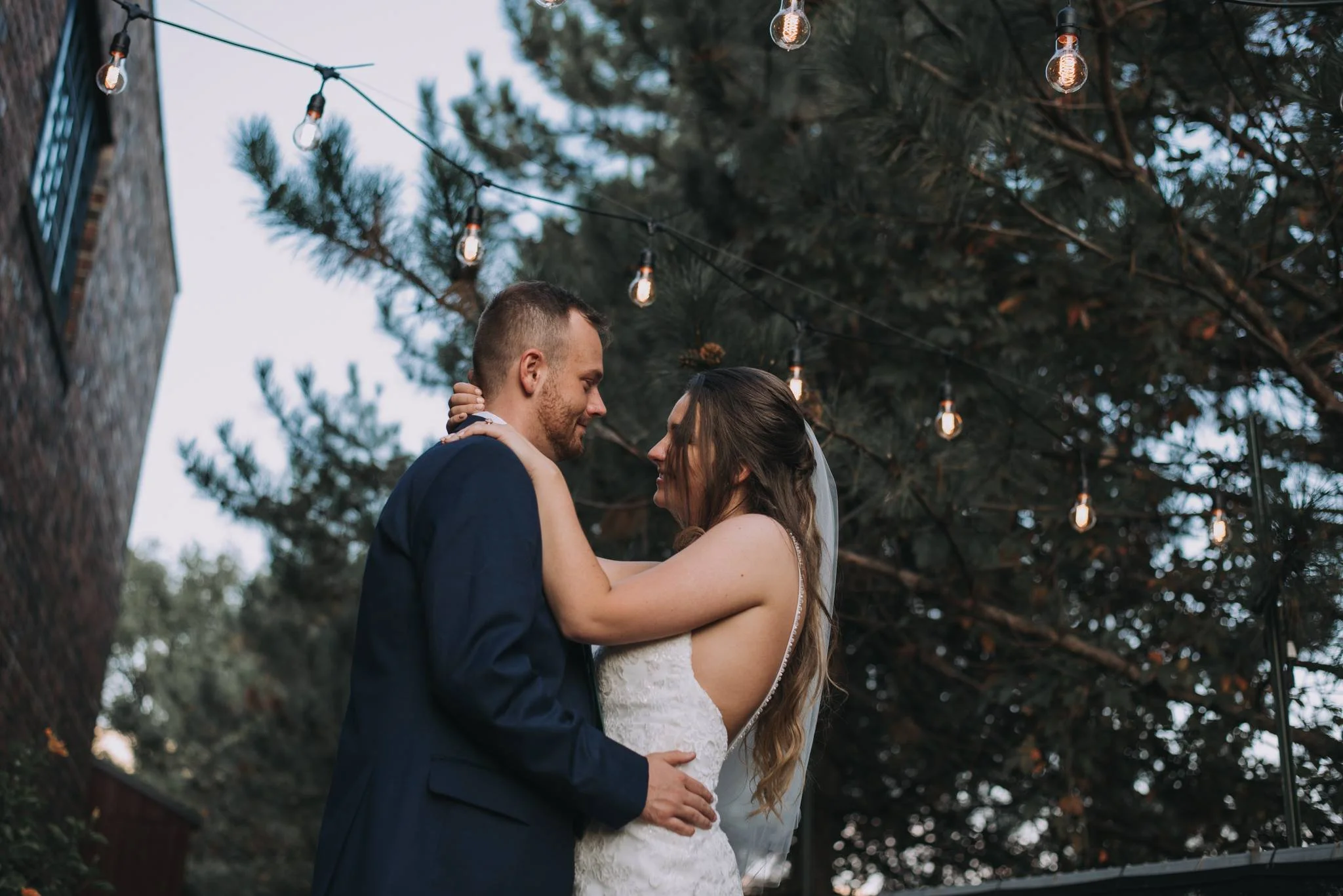 A bride and groom sharing a romantic moment outdoors during their wedding, under hanging string lights with trees in the background.