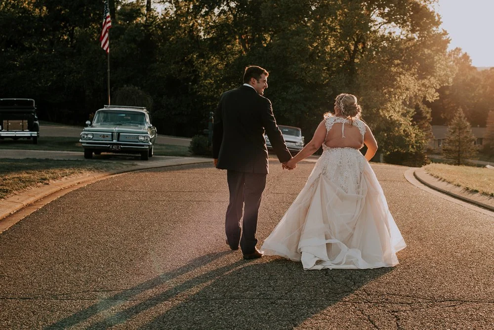 A newlywed couple holding hands and walking away from the camera on a curved street during sunset, with vintage cars and trees in the background.