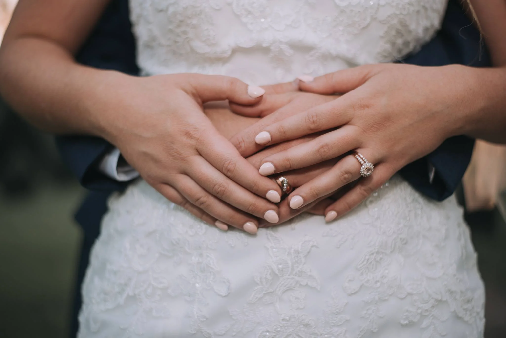 A bride and groom clasp their hands together at their wedding, showing rings and a heart shape formed with their hands, with the bride wearing a wedding dress and the groom in a dark suit.