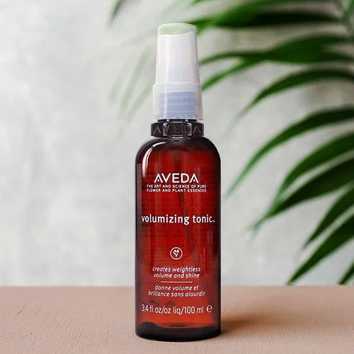 A composite image of a red bottle of Aveda volumizing tonic spray with a white spray cap, placed on a light brown wood surface with green leaves in the background.