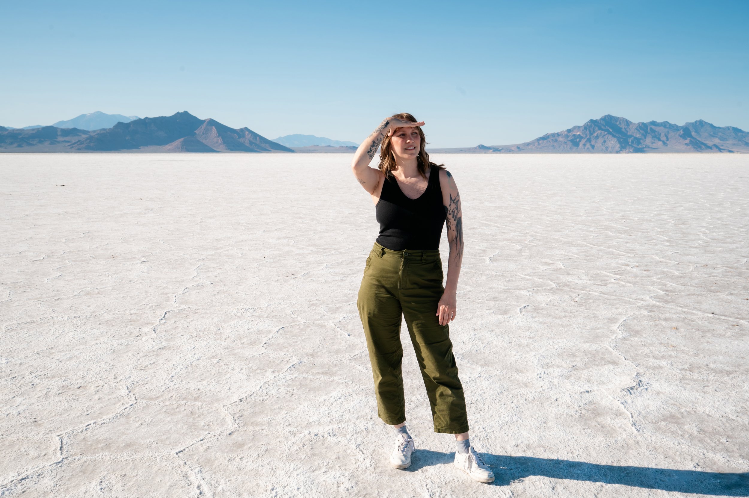 A woman standing on a white salt flat in a desert, with mountains in the background, shielding her eyes from the sun with her hand.