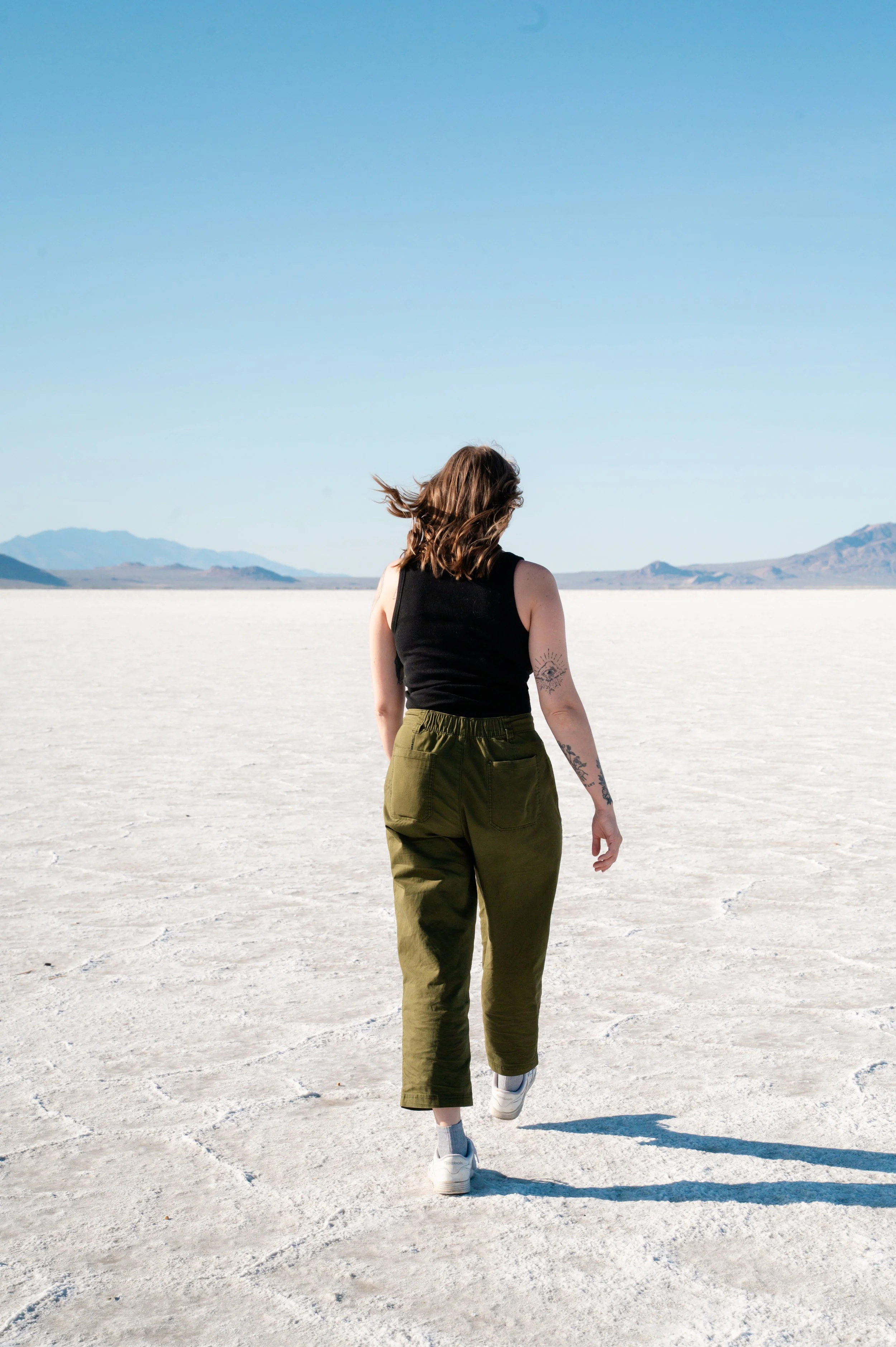 Woman with brown hair in a black sleeveless top and green pants walking in a vast, flat, dry landscape with mountains in the distance under a clear blue sky.