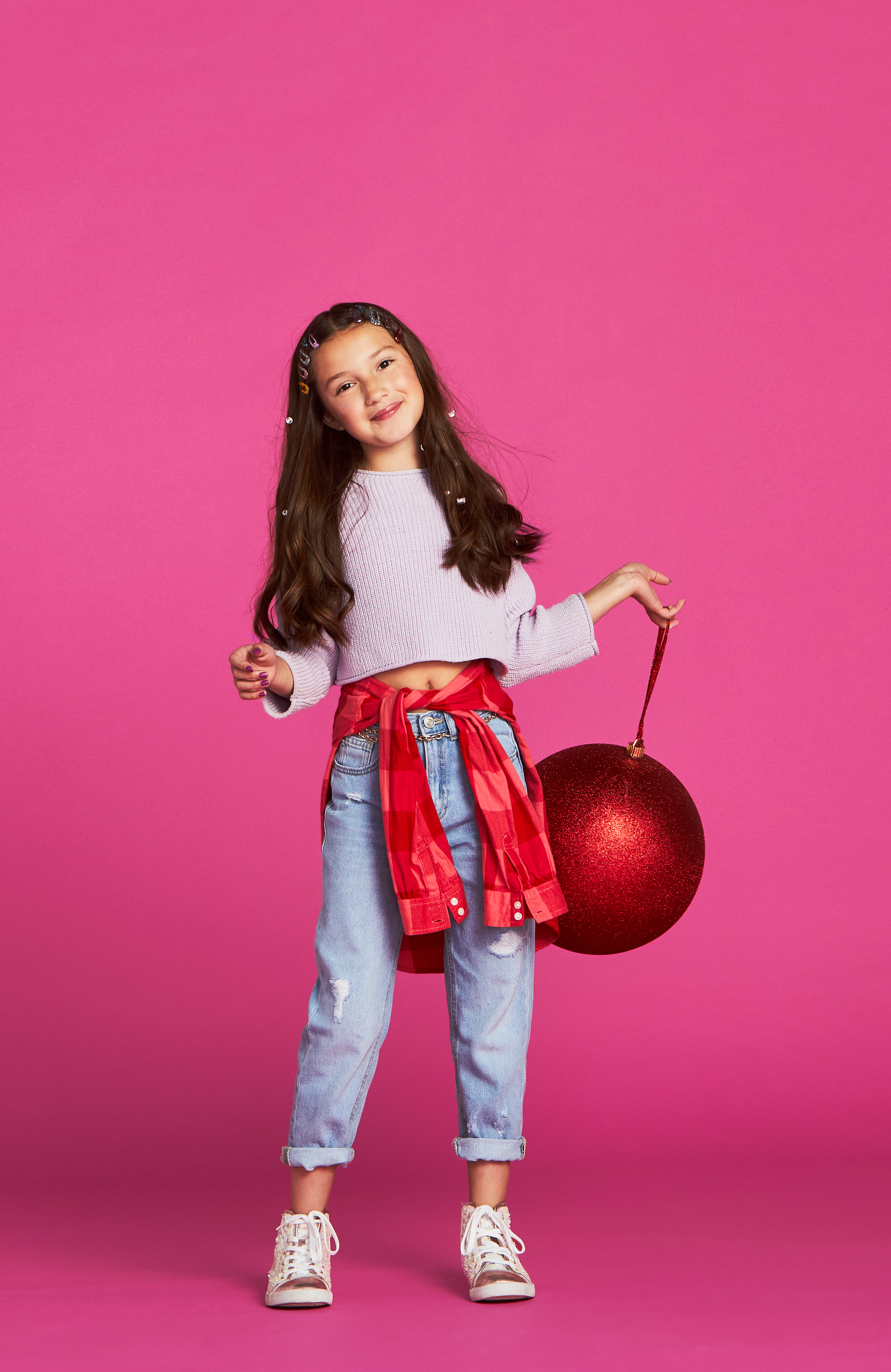 A young girl holding a large red Christmas ornament standing against a pink background.