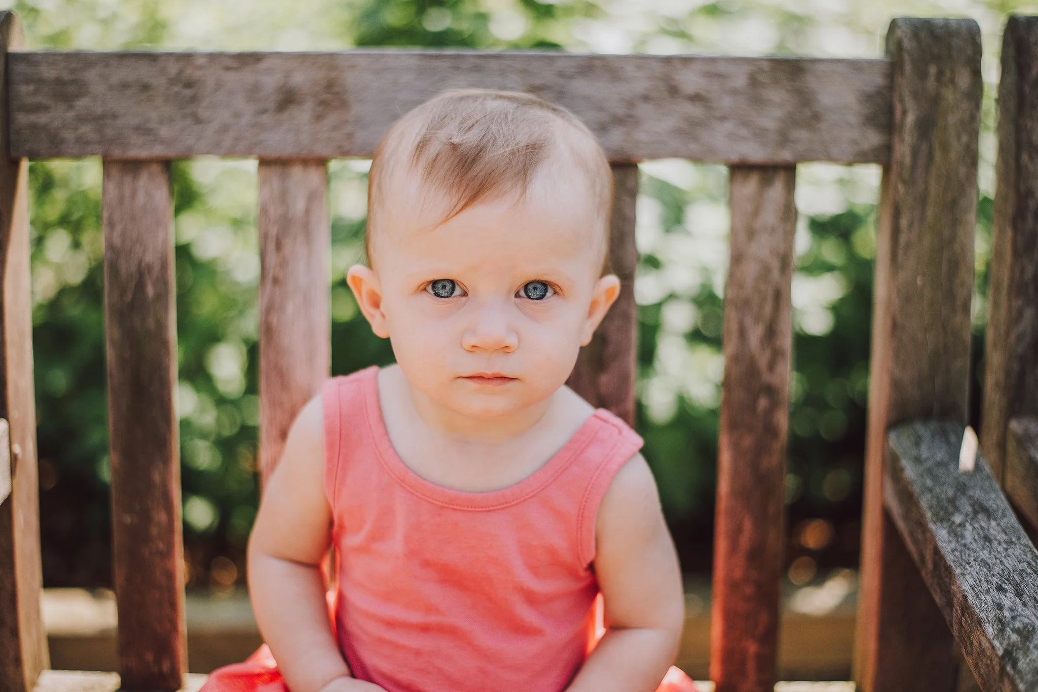A young girl with blue eyes and light hair, wearing a pink sleeveless top, sitting on a wooden bench outdoors with green foliage in the background.