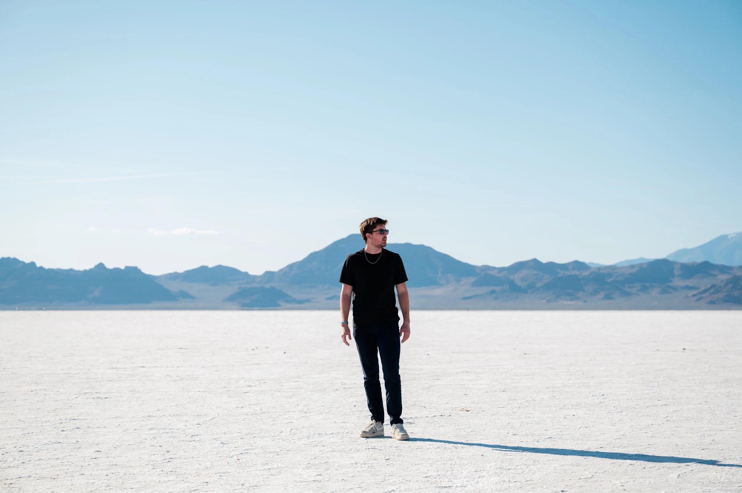 A young man walks across a vast, flat, salt flat landscape with mountains in the background under a clear blue sky.