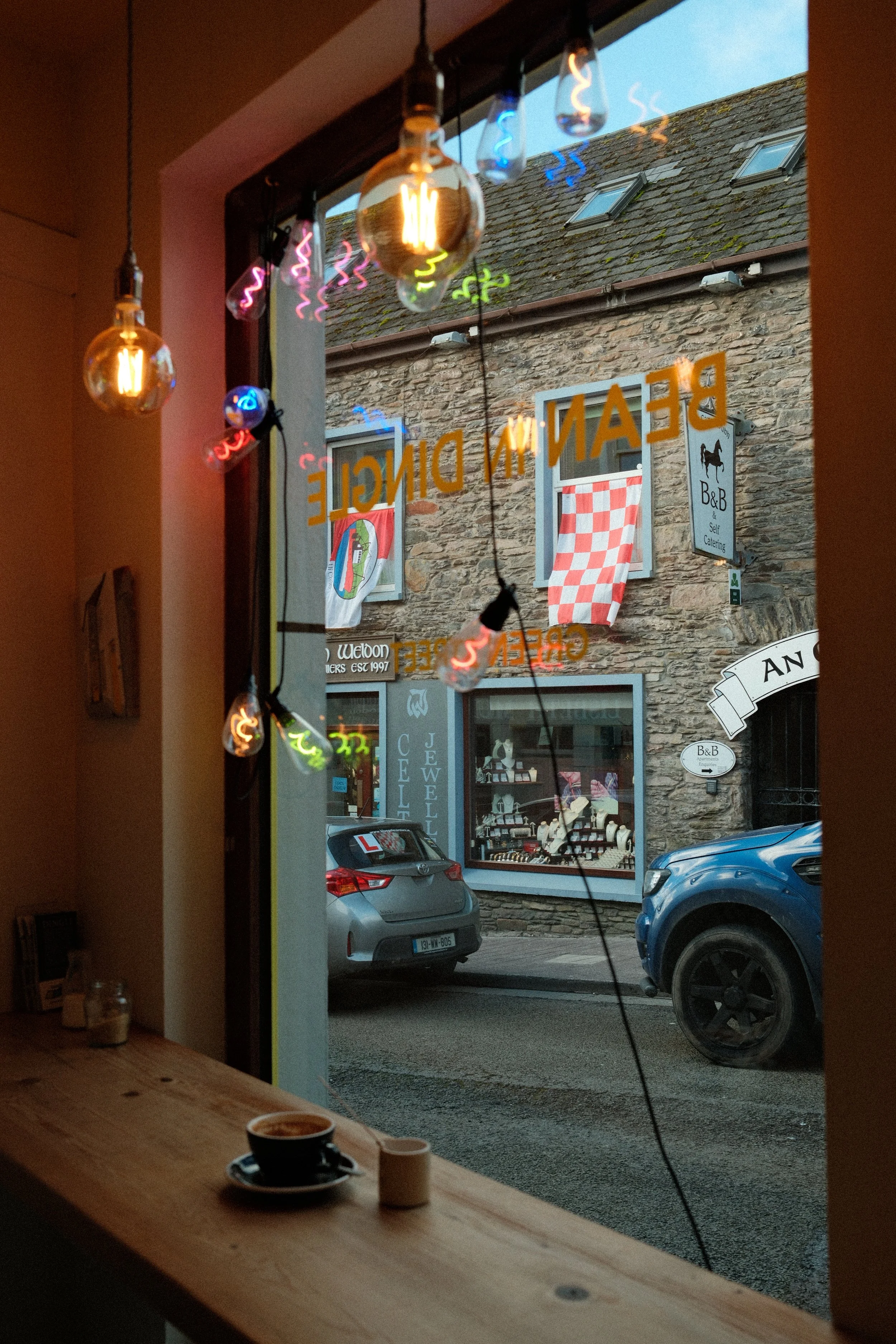 Inside a cafe, looking out a window with hanging Edison-style light bulbs, a coffee cup on the counter, and cars parked outside on a street with stone buildings and signs.