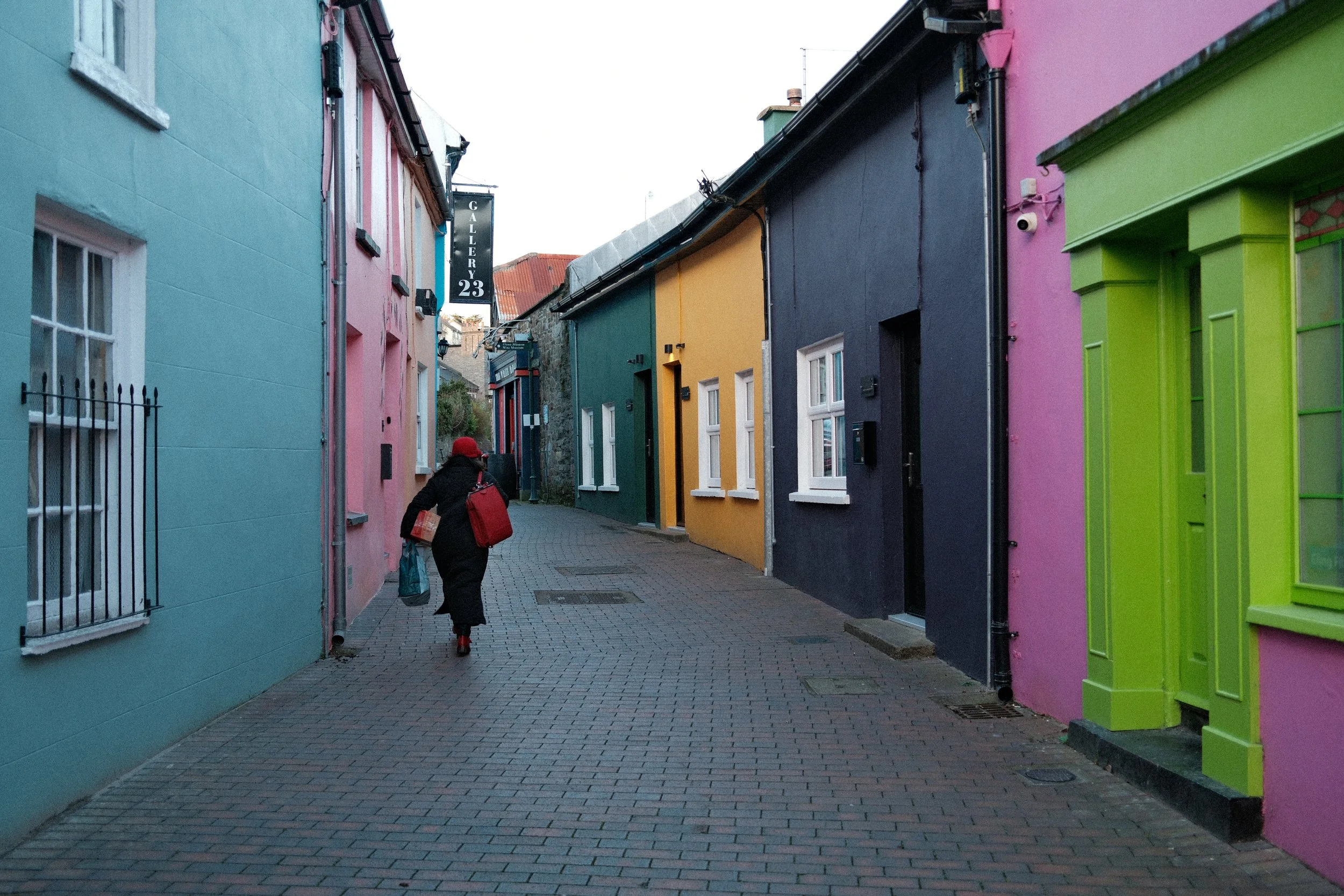 A narrow street with colorful buildings painted in pastel shades including blue, pink, green, yellow, and dark blue. A person with a red hat and black coat walks away carrying shopping bags.