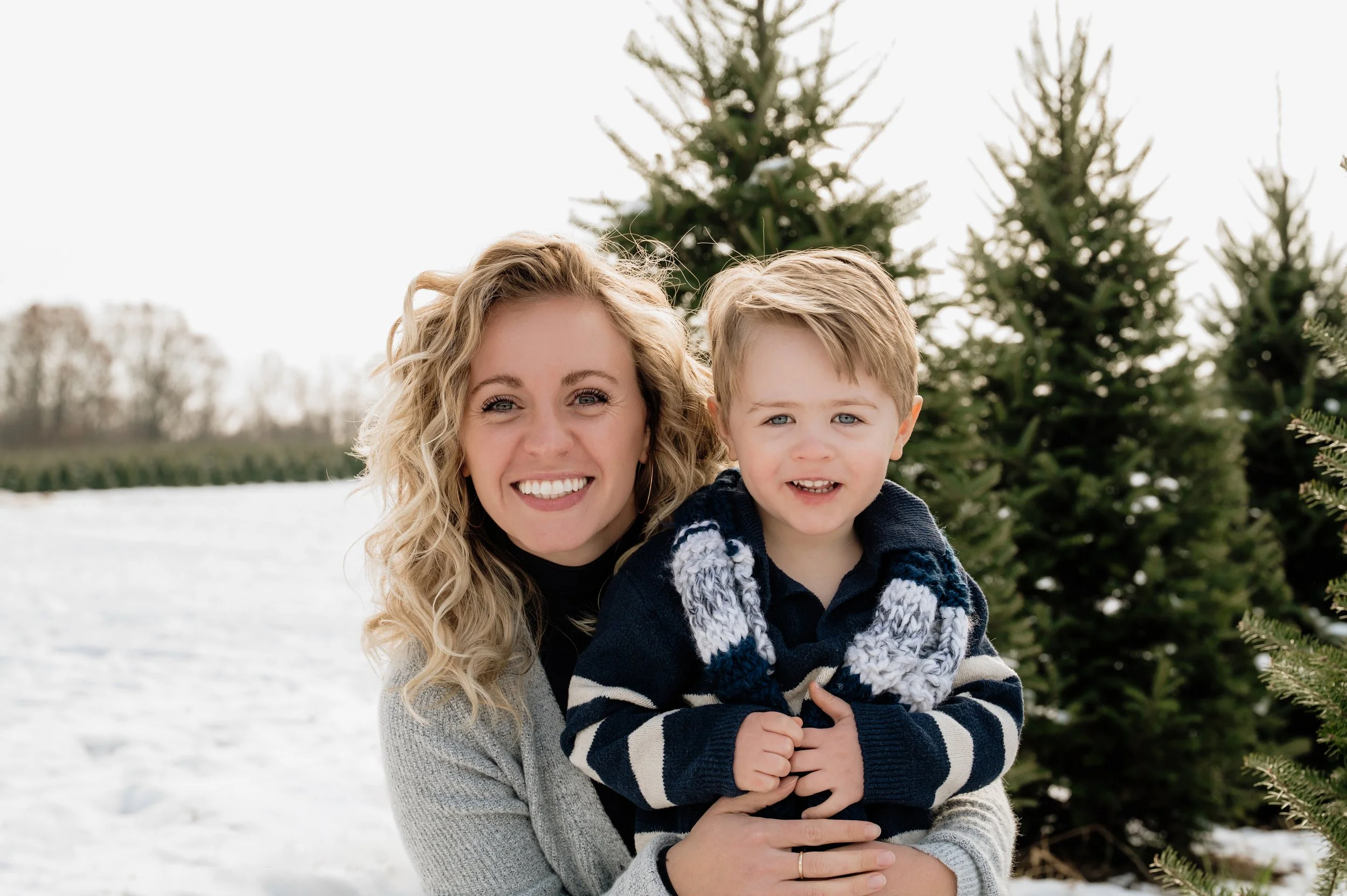 A smiling woman with curly blonde hair holding a young boy with short blonde hair outdoors in front of evergreen trees and snow.