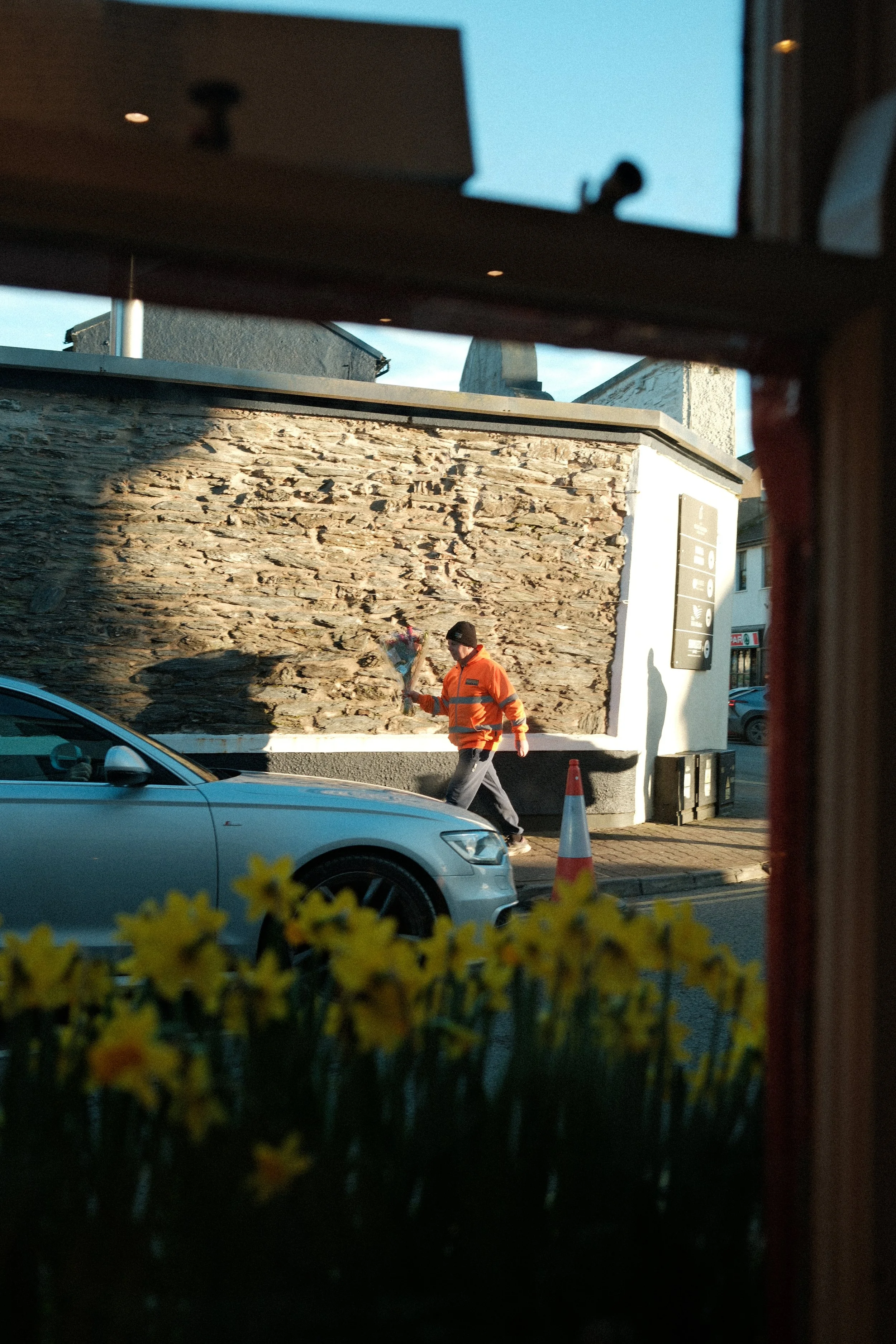 A man in an orange reflective jacket walking past a wall, seen through a window with yellow flowers at the bottom and part of a table or window frame at the top. A silver car is parked nearby.