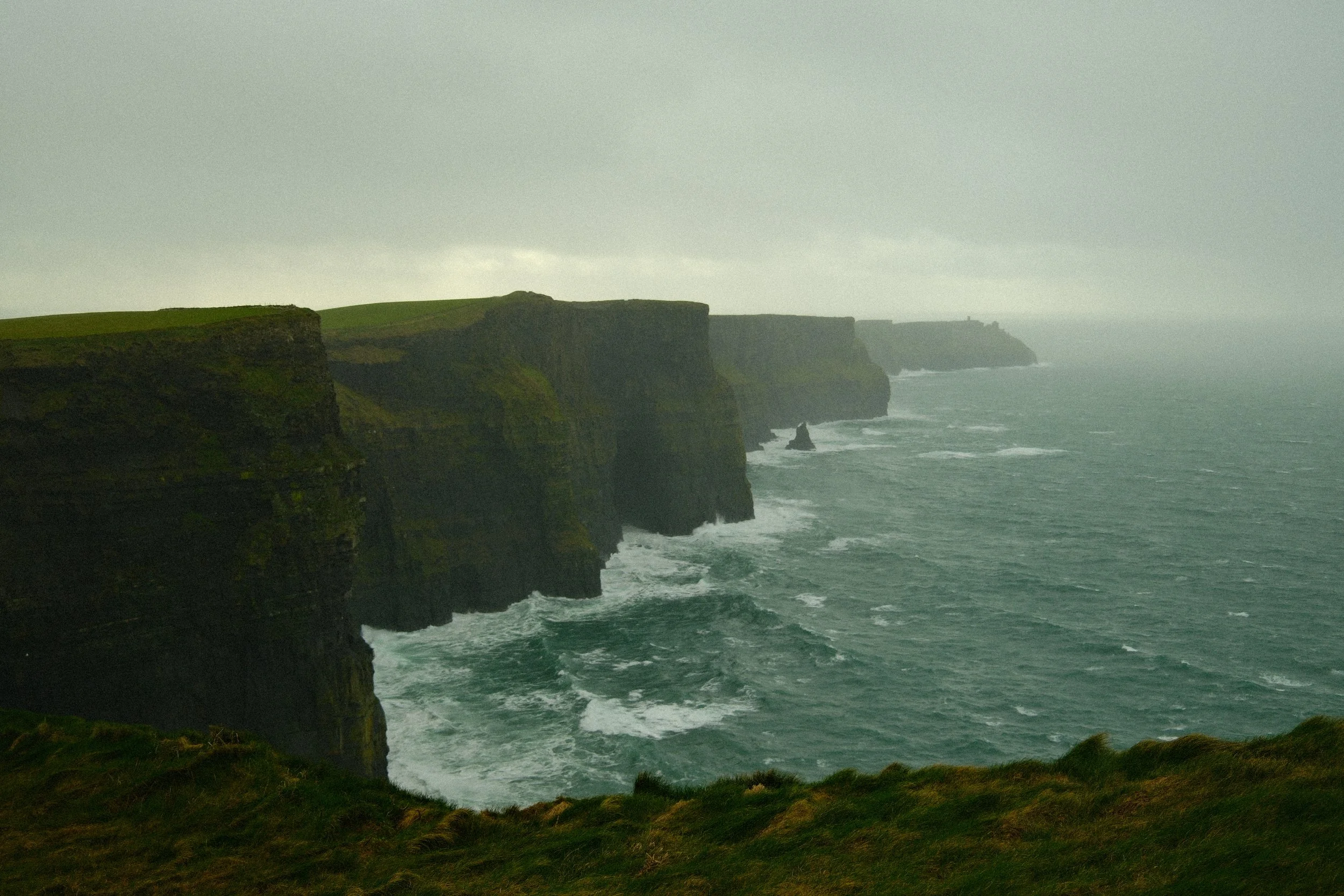 Cliffs along a rough sea under cloudy sky with a grassy foreground.