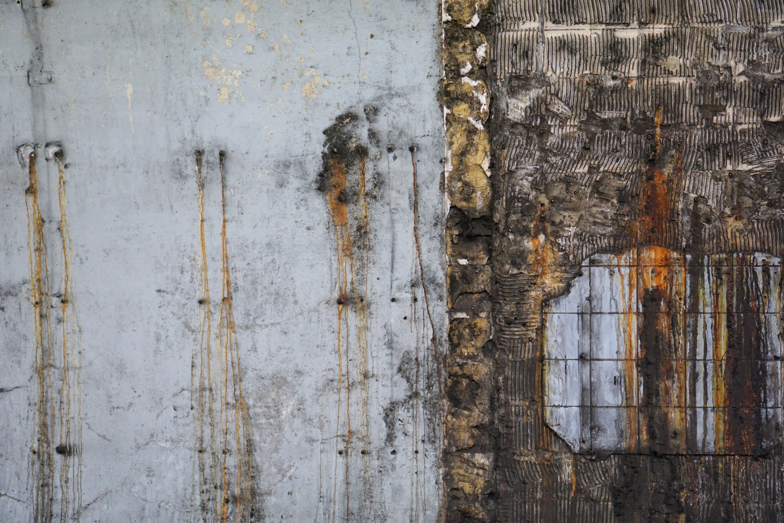 A weathered wall with peeling paint and rust stains, showing sections of concrete and brick layers.