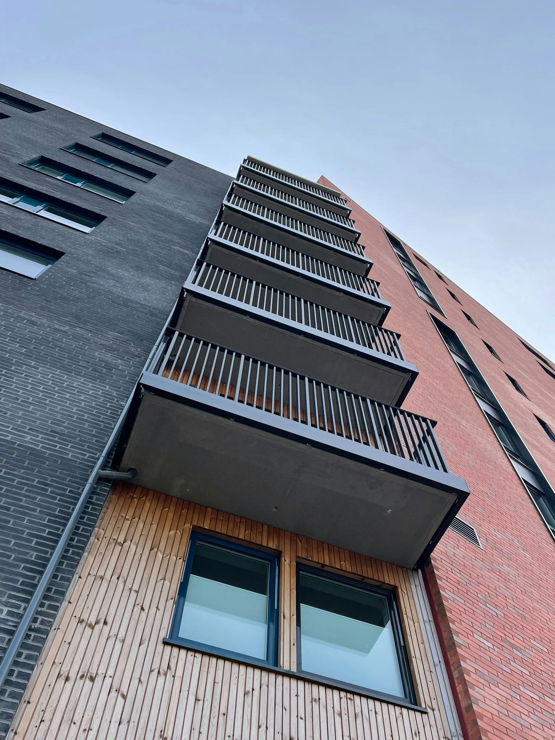 Low-angle view of a modern multi-story building with brick, dark gray, and wood exterior, featuring balconies with black railings and large windows, under a clear blue sky.