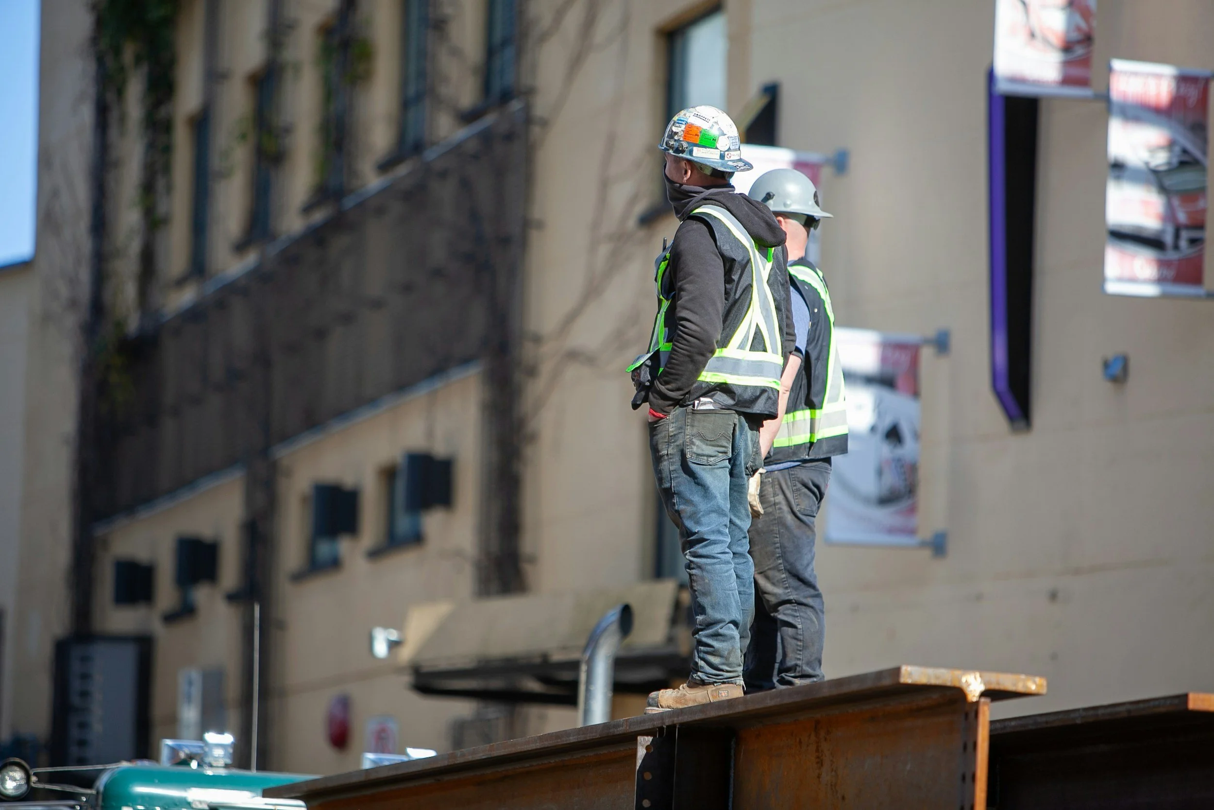 Two construction workers in safety vests and helmets standing on a platform outside a building under construction.
