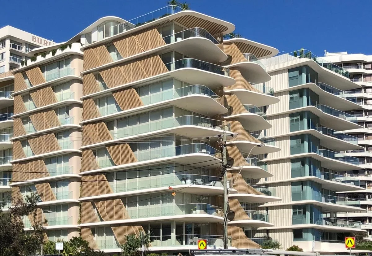 Modern multi-story residential building with curved balconies and wooden window shutters, set against a bright blue sky.