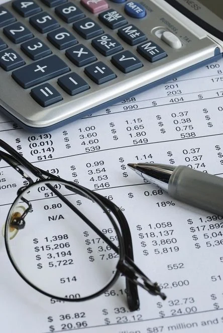 Close-up of a calculator, a pair of eyeglasses, and financial documents with printed numbers and prices, along with a pen resting on the paper.