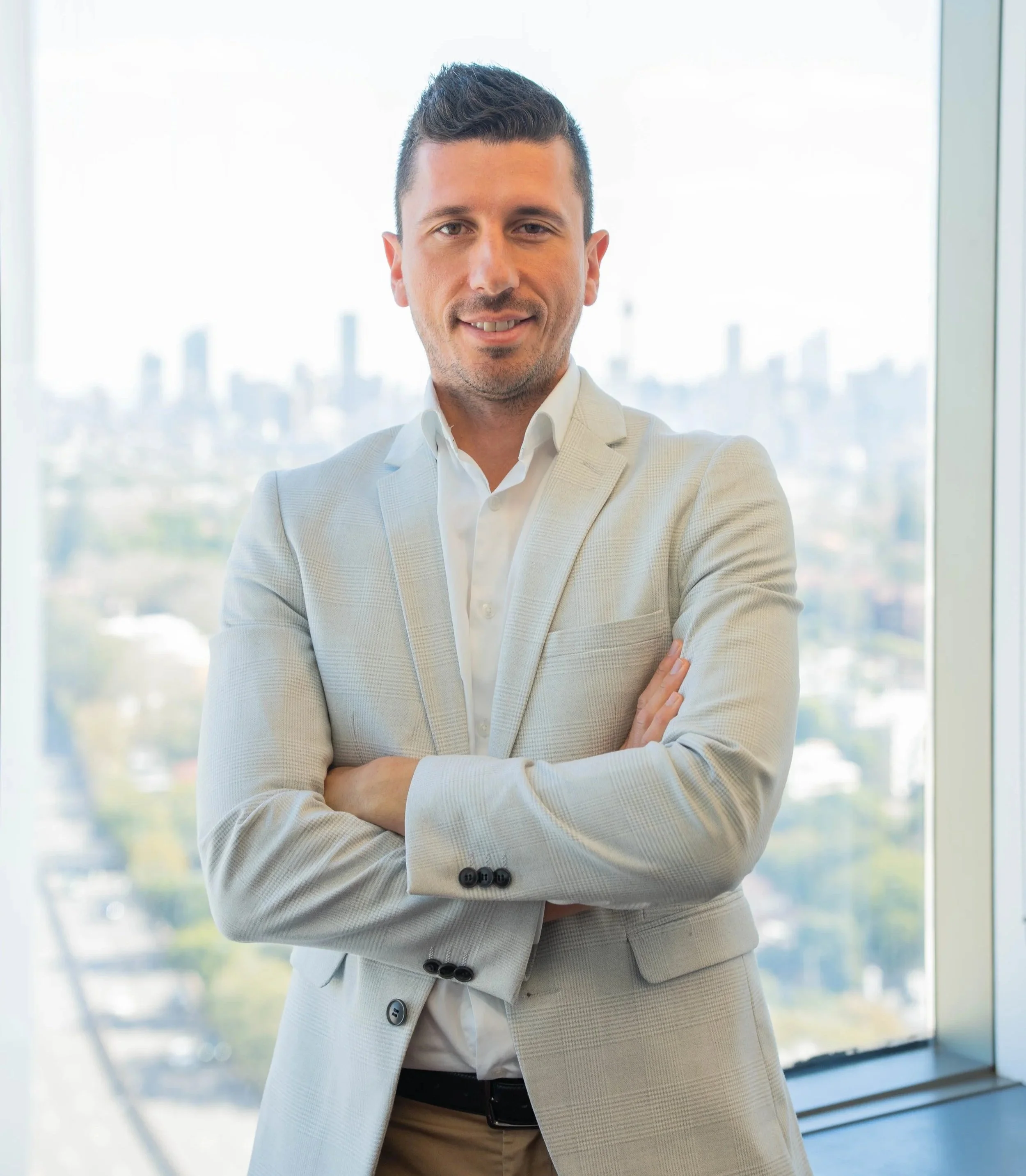 Professional man in a light-colored suit with crossed arms, standing indoors near large windows with cityscape in the background.