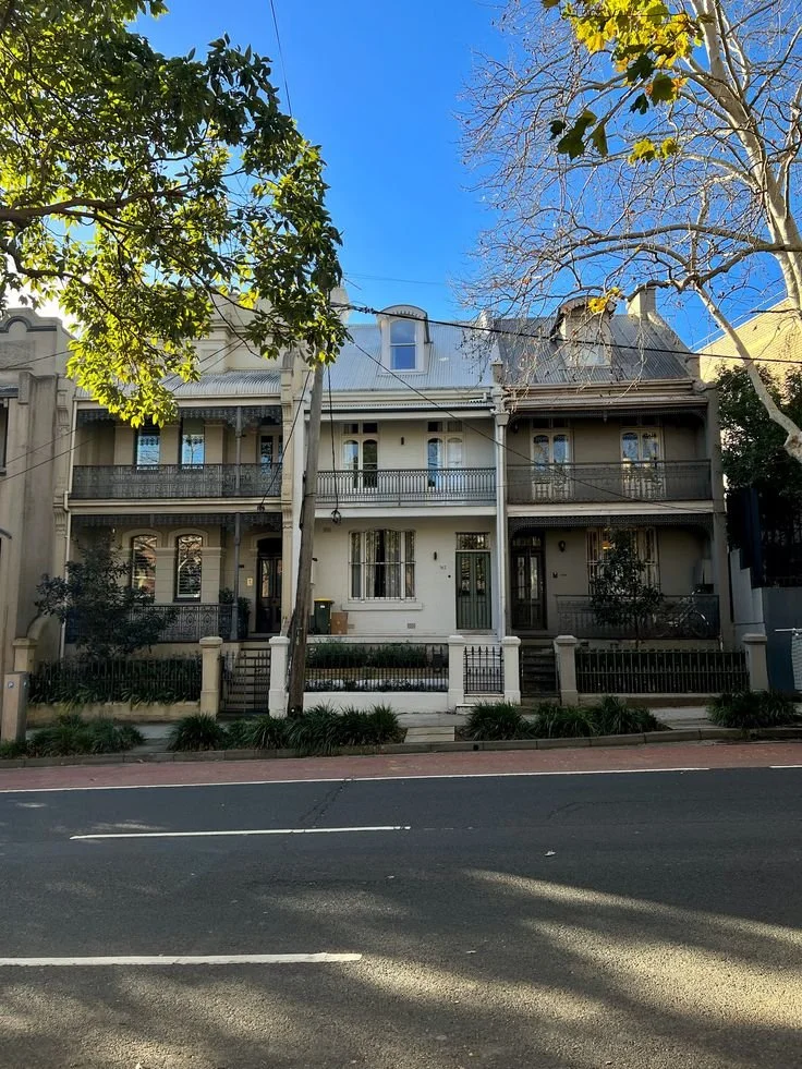 A row of three-story Victorian-style townhouses with front porches, iron railings, and large windows. The street in front has a bike lane and some parked cars, with trees and clear blue sky in the background.