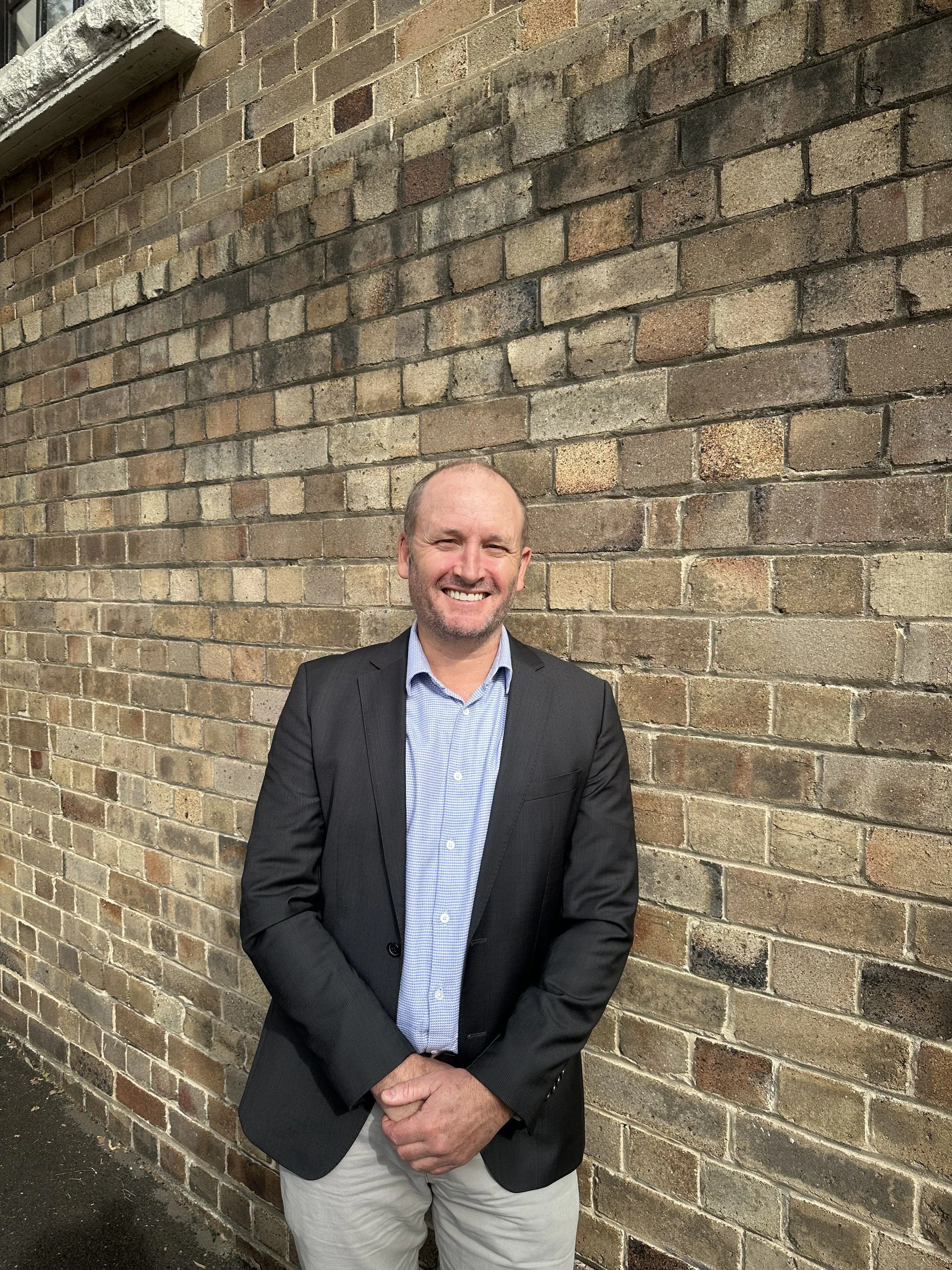 A man smiling, standing in front of a brick wall, wearing a suit jacket and light-colored pants.