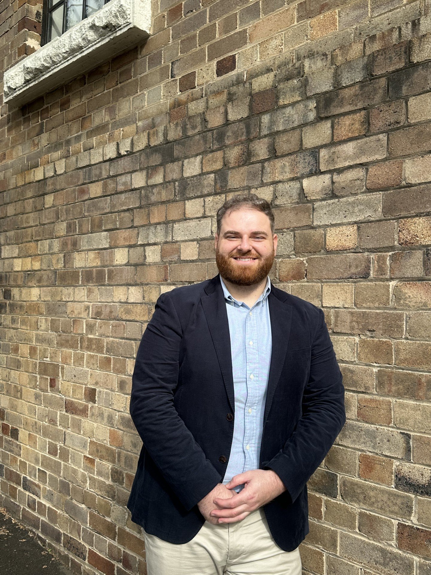 A man with a beard and light hair wearing a dark blazer, light blue shirt, and light khaki pants standing in front of a brick wall.