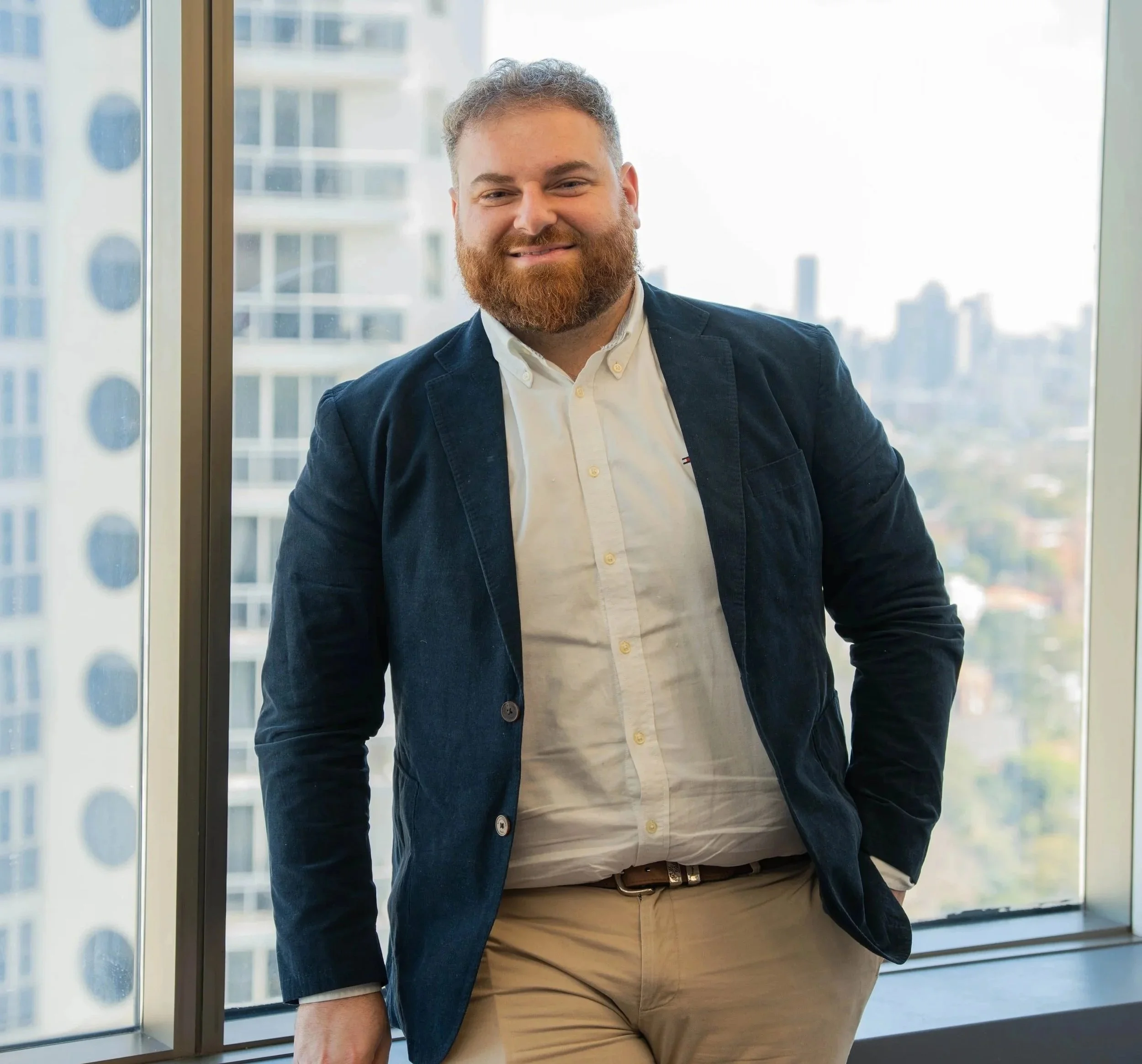 A man with curly hair and a beard smiling, dressed in a white shirt and blue blazer, standing near a large window with city buildings in the background.
