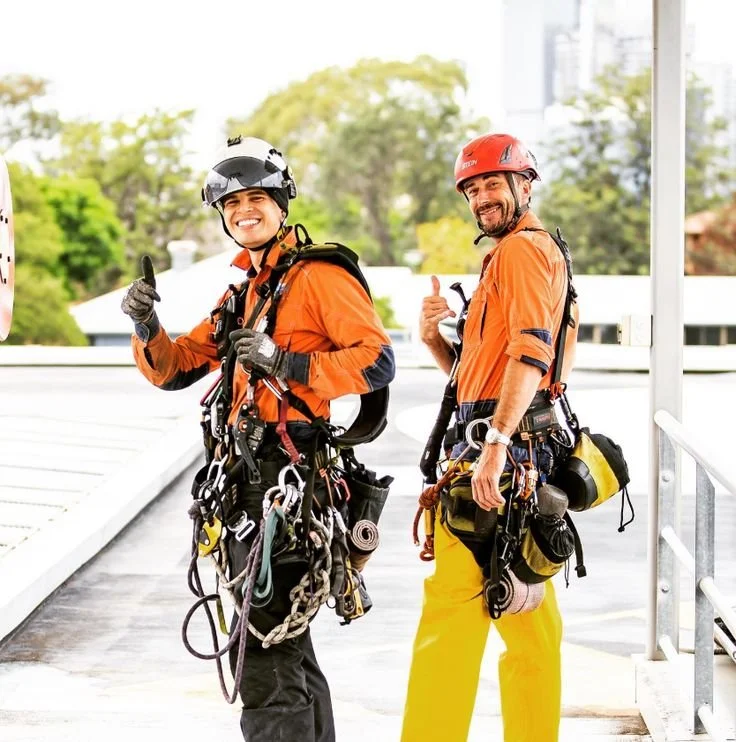 Two smiling male climbers in safety gear, giving thumbs up, standing outdoors on a rooftop or balcony.