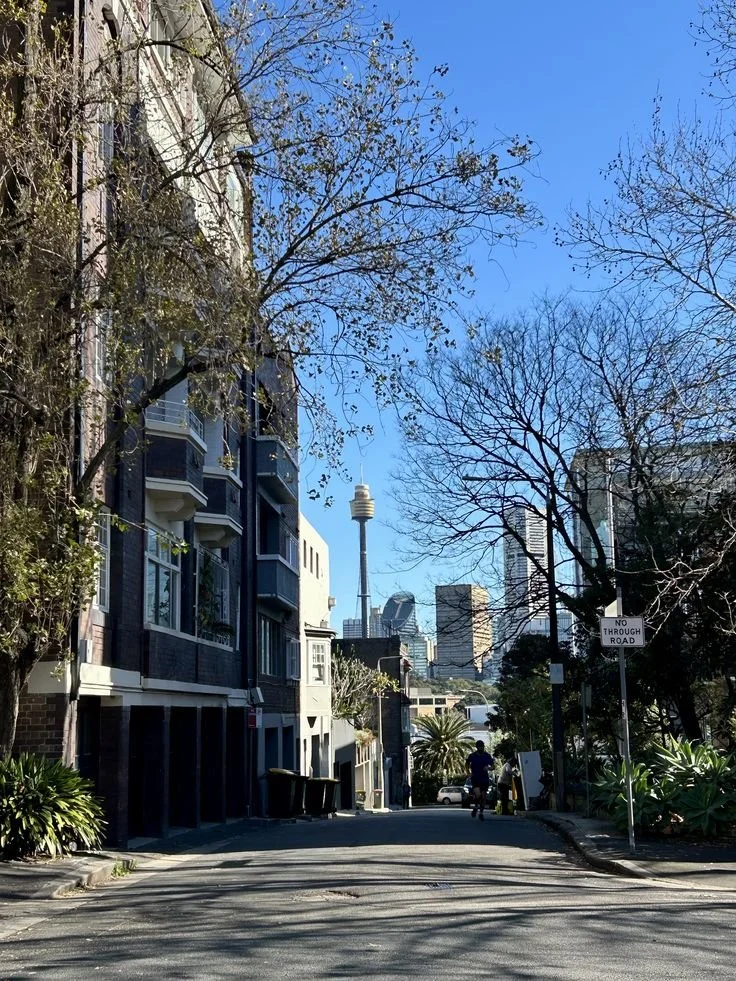 A quiet city residential street with apartment buildings on the left, trees with sparse leaves, and a skyline featuring the Sky Tower in the distance, under a clear blue sky.