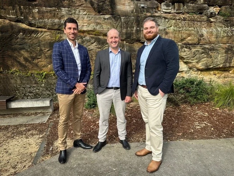 Three men in business casual attire standing outdoors on a concrete path, with a rocky and leafy background.