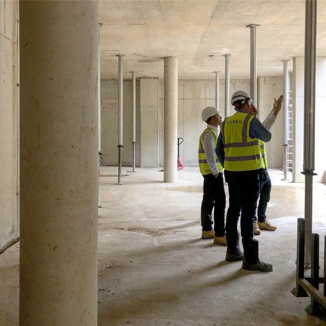 Three construction workers in safety helmets and high-visibility vests are standing inside a building under construction, looking at the space and one is talking on a phone.