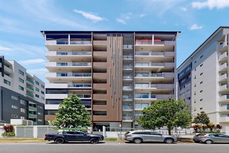 Six-story modern apartment building with balconies on each floor, flanked by similar buildings, with trees and parked cars in front under a blue sky.