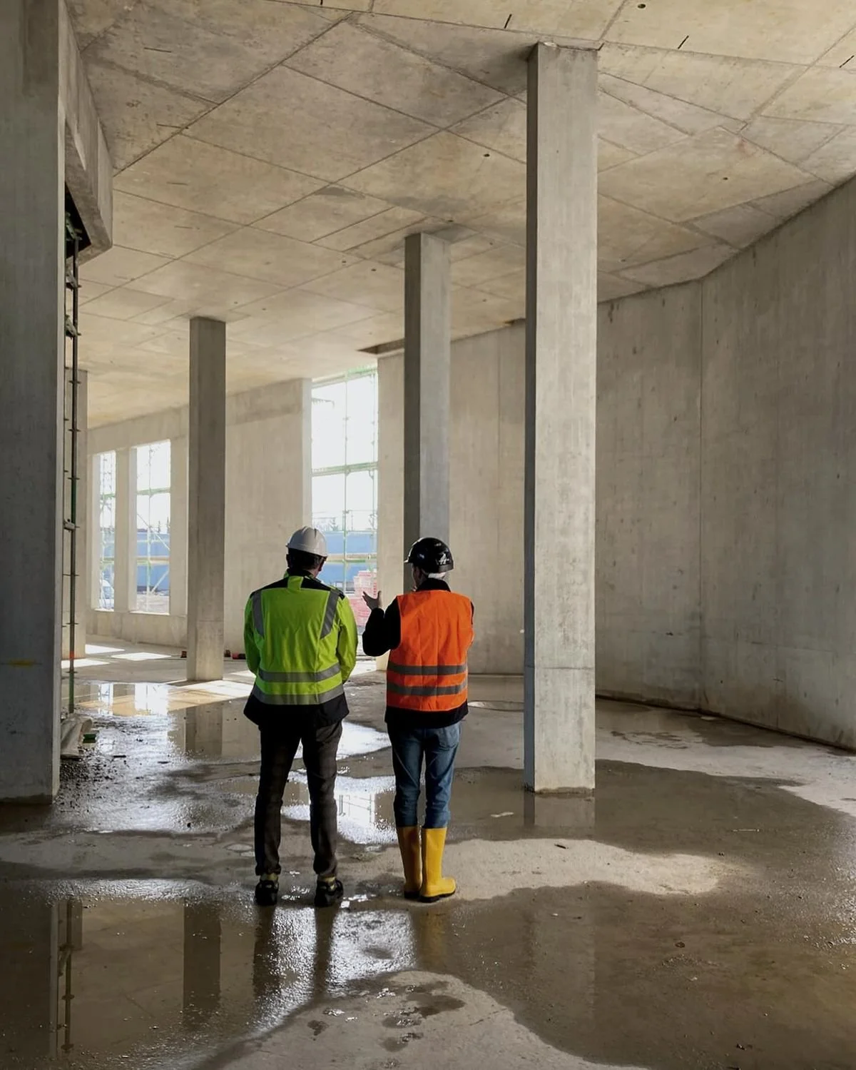 Two construction workers in safety vests and helmets standing inside a large unfinished building with concrete walls and columns, wet floor, and puddles of water.