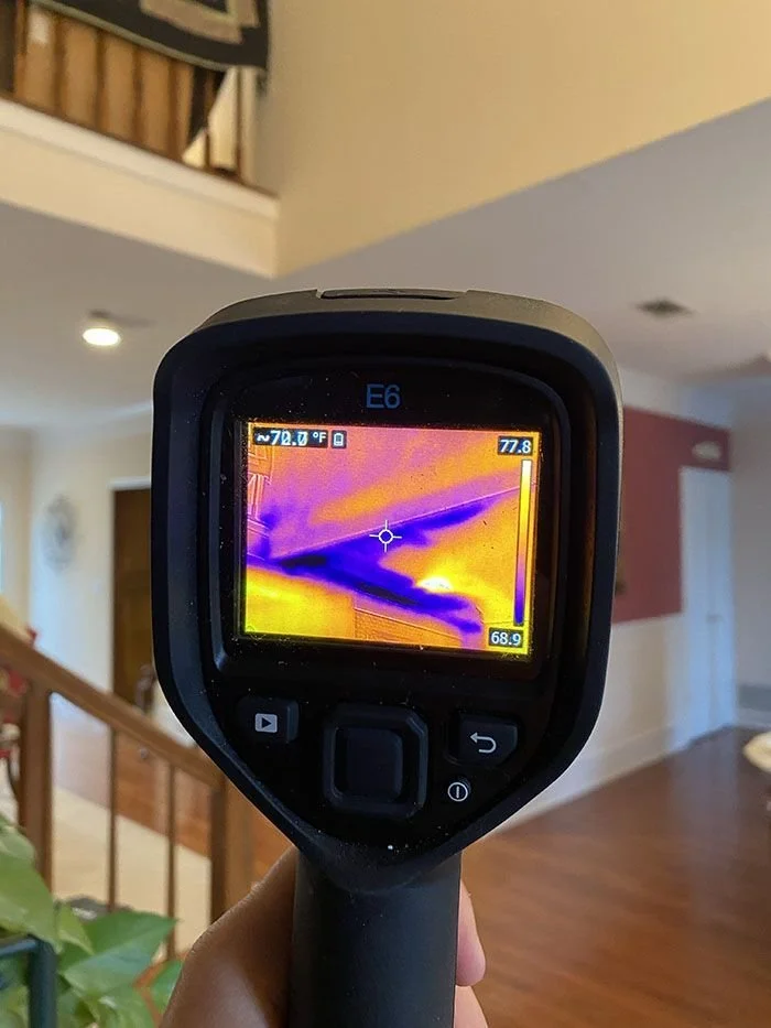 Handheld infrared thermal camera showing a thermal image of the ceiling in a room, with various colors indicating temperature differences.