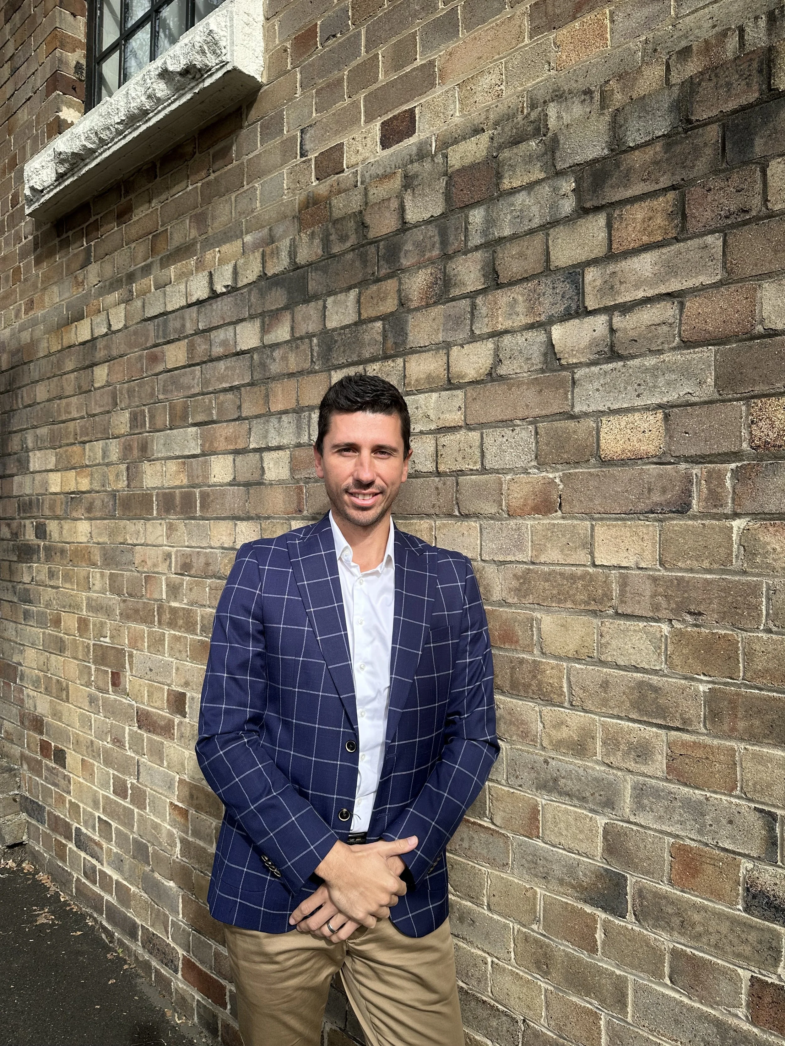 A man with short dark hair and light skin, dressed in a navy blue plaid blazer and beige pants, standing in front of a brick wall with a large window at the top left corner, smiling at the camera.
