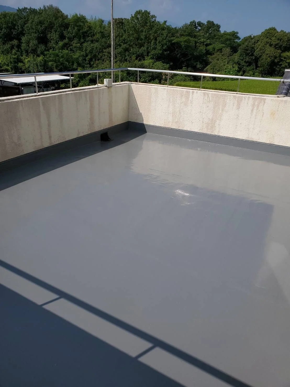 Empty rooftop terrace with fresh gray waterproof coating and a metal railing, overlooking green trees and a blue sky.