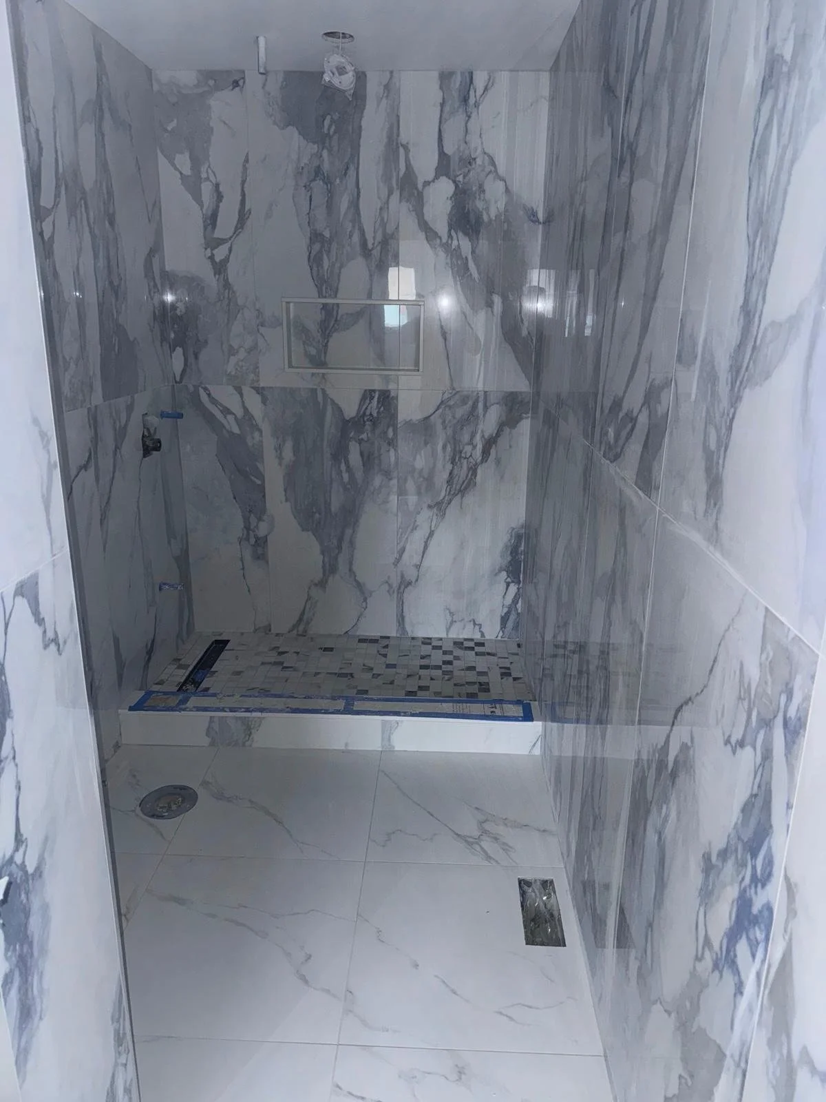Empty walk-in shower with marble walls and floor, a small built-in niche, a floor drain, and a linear drain at the entrance, under construction.