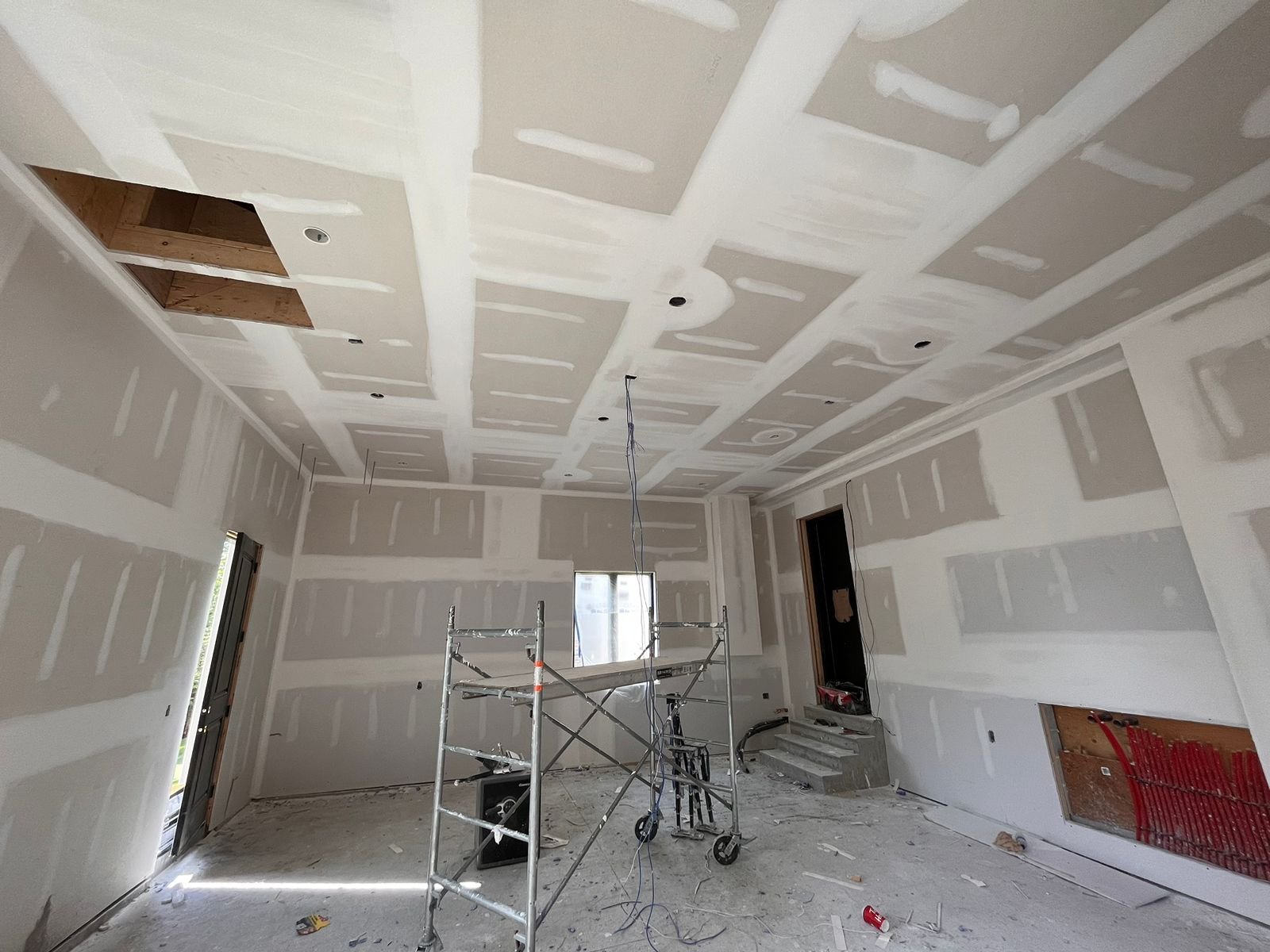 Interior room under construction with drywall installation, a scaffold standing in the center, exposed ceiling with cutouts, and construction tools and debris on the floor.
