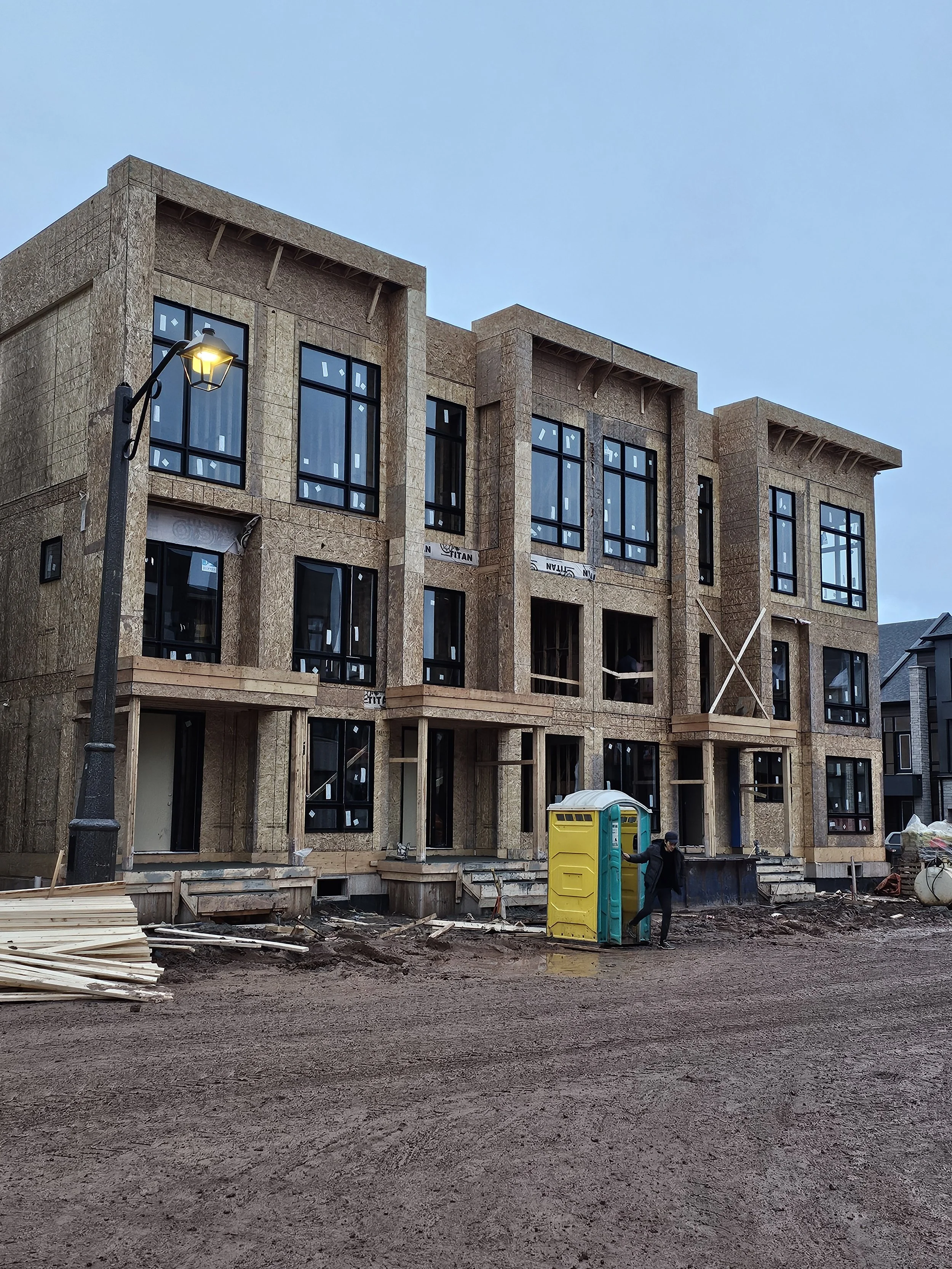 Under construction multi-story building with large windows, partially built, on muddy ground, with a street lamp and a portable toilet in the foreground, and a person standing beside the toilet.