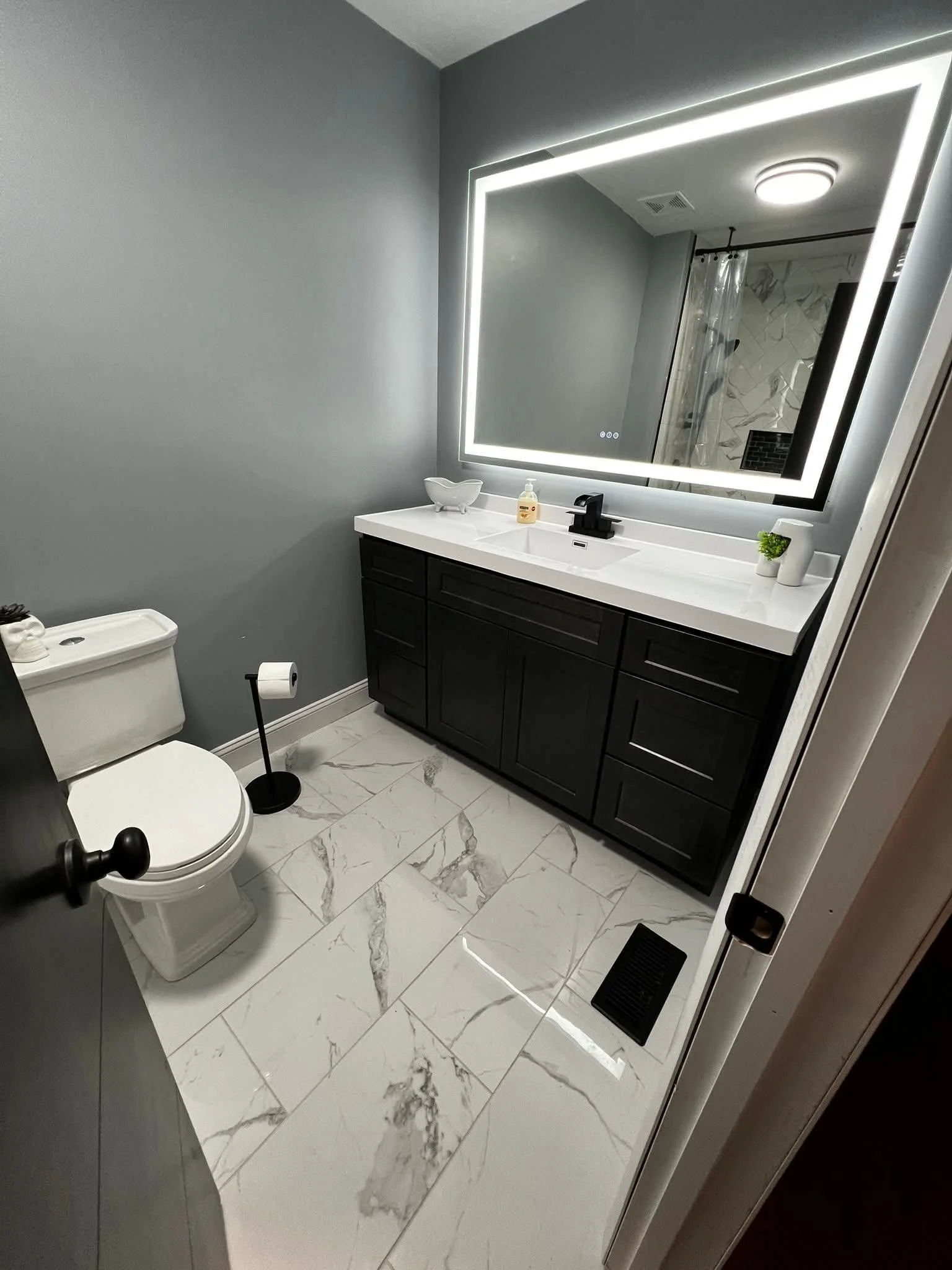A modern bathroom with a white toilet, black vanity with a white countertop, large illuminated mirror, and marble tile flooring.