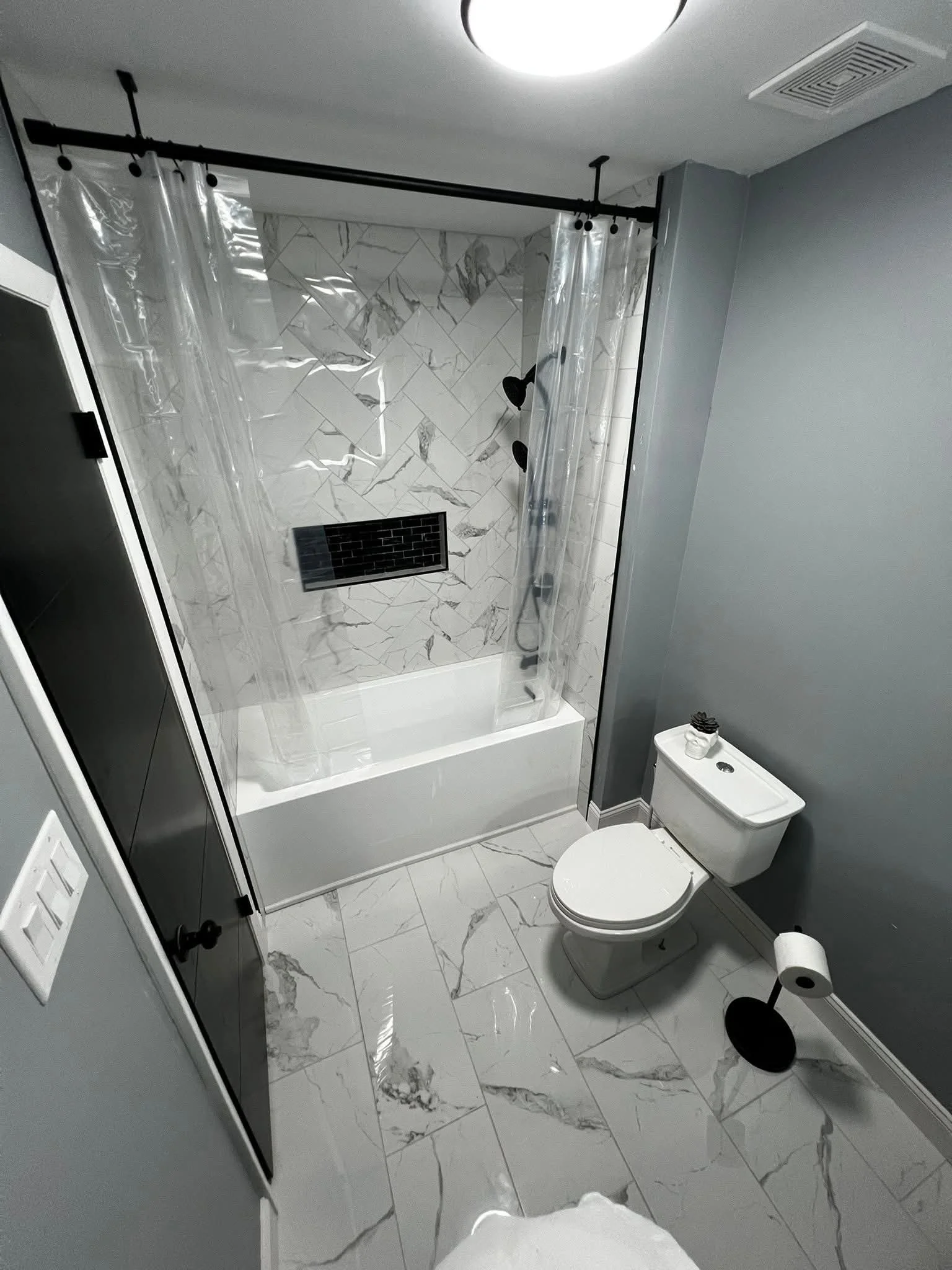 Modern bathroom with gray walls and marble floor and shower wall. White bathtub with a clear shower curtain, black shower fixtures, and a black vent in the tiled shower. White toilet with black-and-white decorative item on top and a black toilet paper holder with a white roll. Ceiling light and vent.