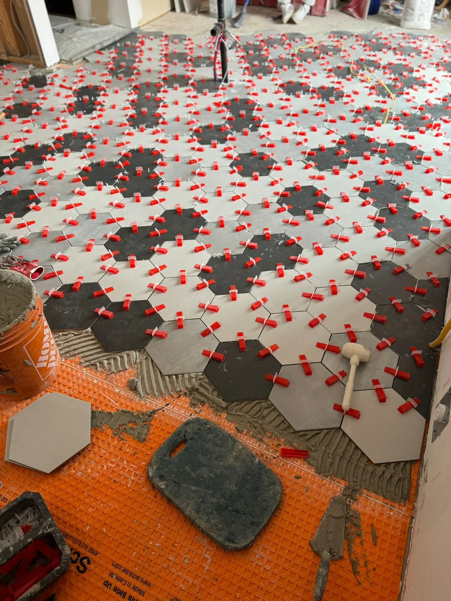 Construction site with hexagonal tiles being installed. Some tiles are black and some are white. Red spacers are placed between the tiles to ensure proper gaps. Tools and construction materials are visible around the floor.