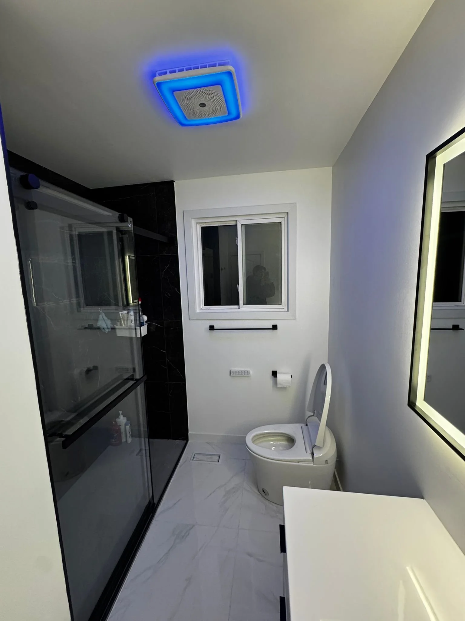 Modern bathroom with white walls, a black and glass shower enclosure, a window, a toilet, a large illuminated mirror, and a white floor with subtle marbling.