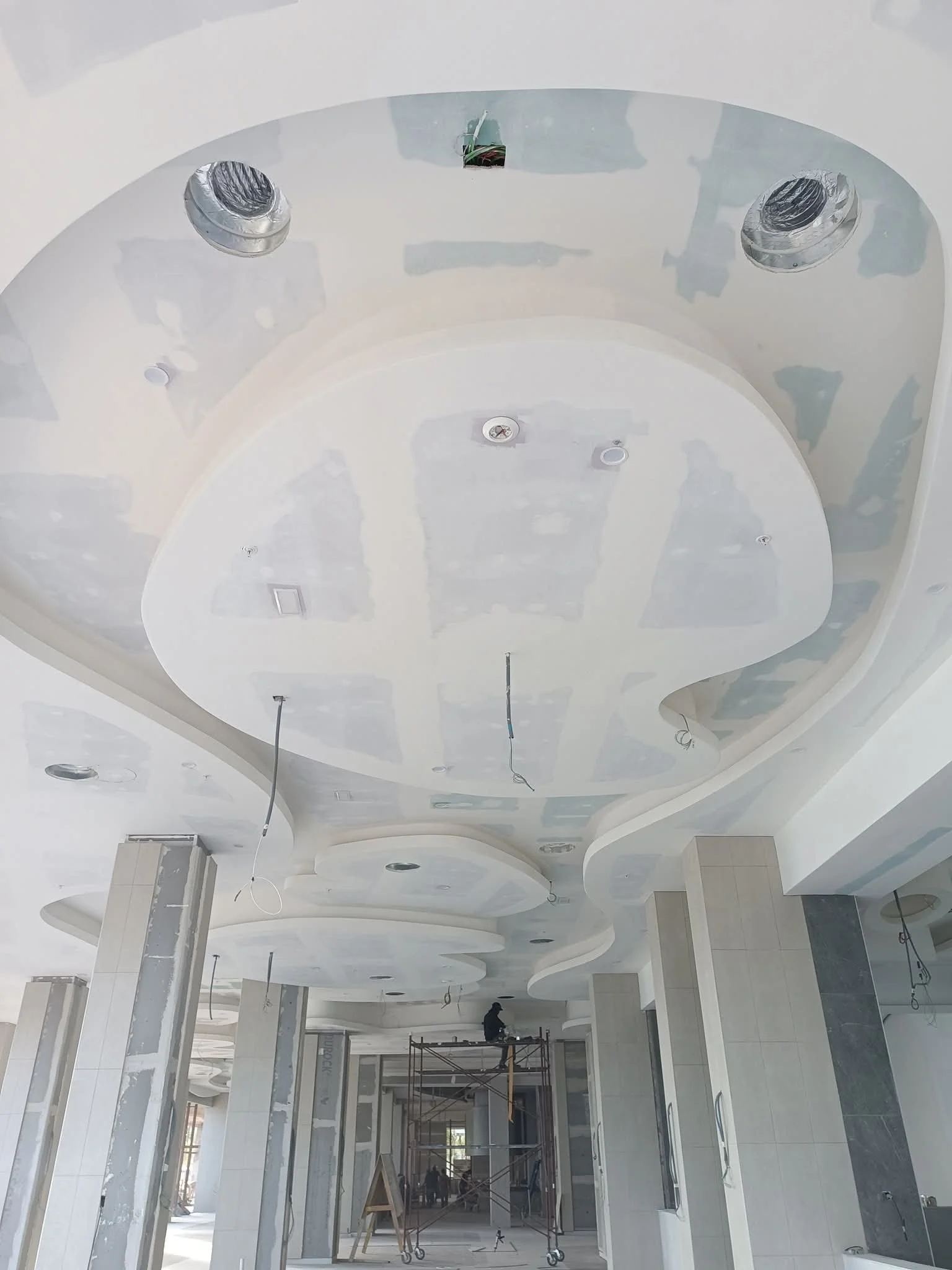 Ceiling construction with recessed lighting fixtures and a worker on scaffolding, in a large room with tiled walls.