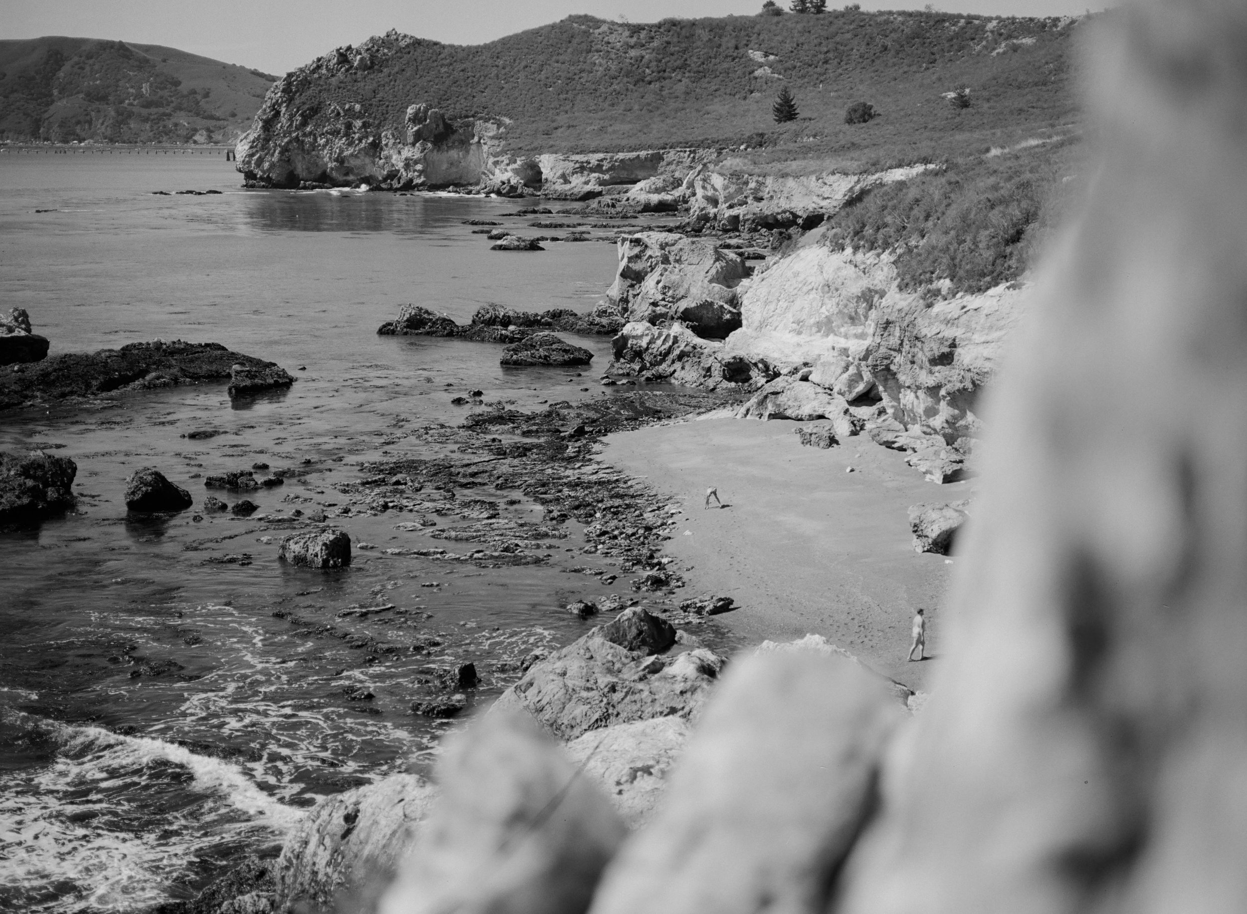 Black and white photo of a rocky beach with cliffs and ocean waves, two people walking along the shoreline, hills in the background.