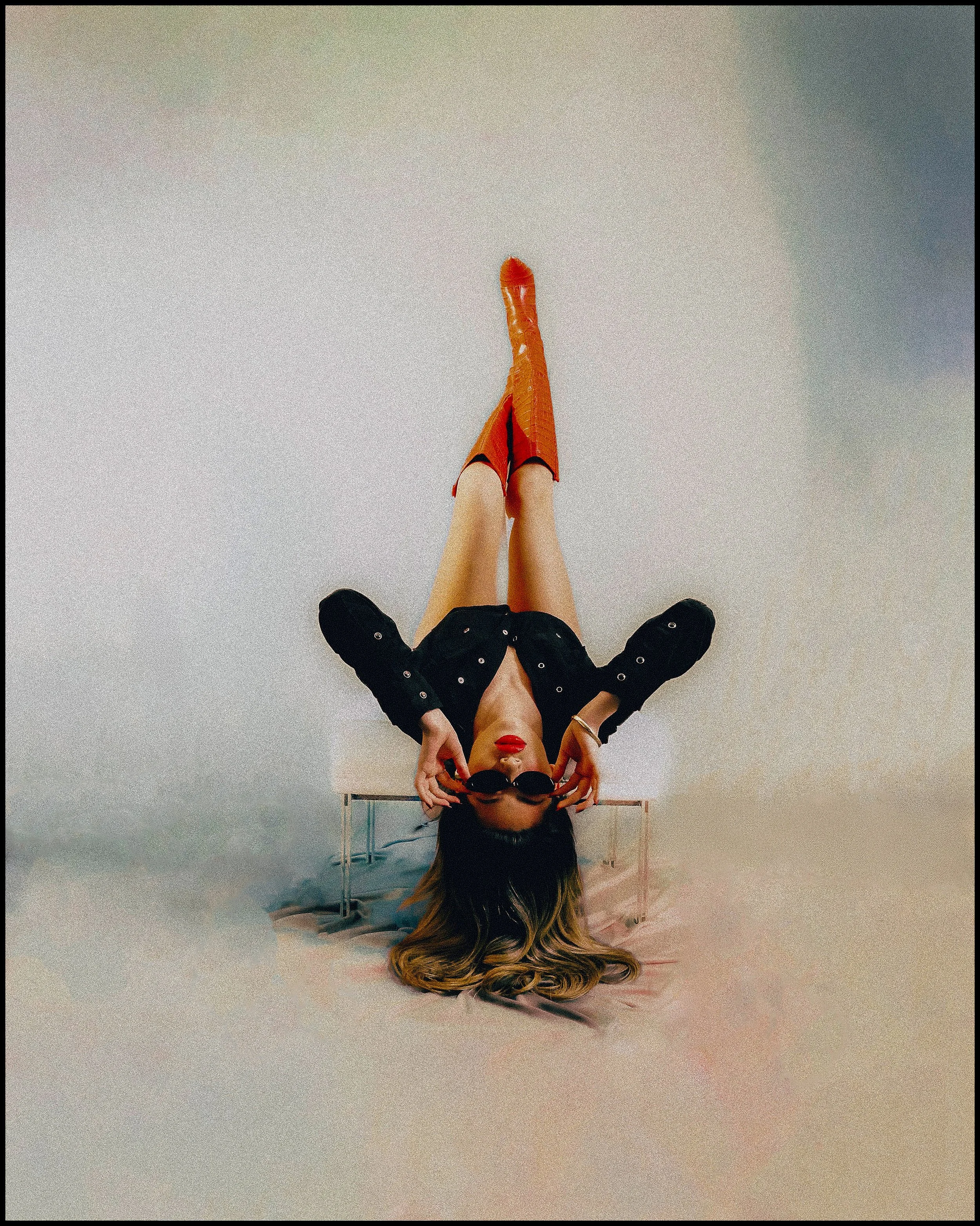 A woman with long hair lying upside down on a small bench, wearing red high heels, black shorts, a black jacket, and sunglasses, against a plain background.