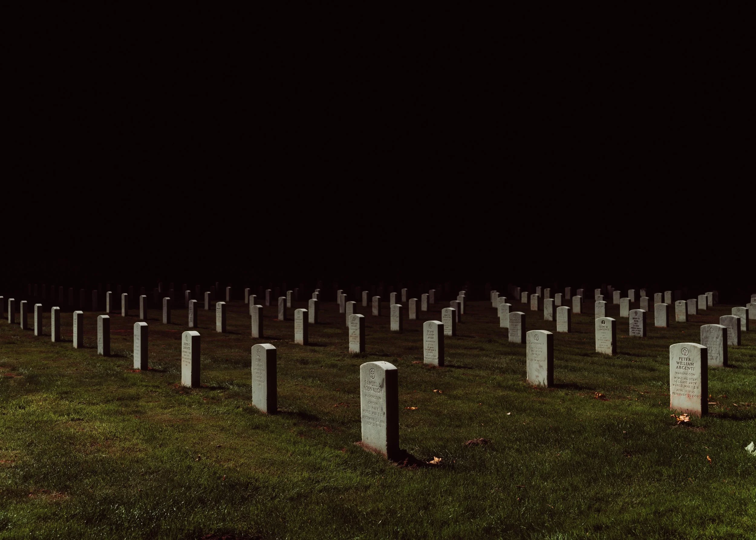 A dark night scene of rows of white gravestones in a cemetery with grass, under a pitch-black sky.