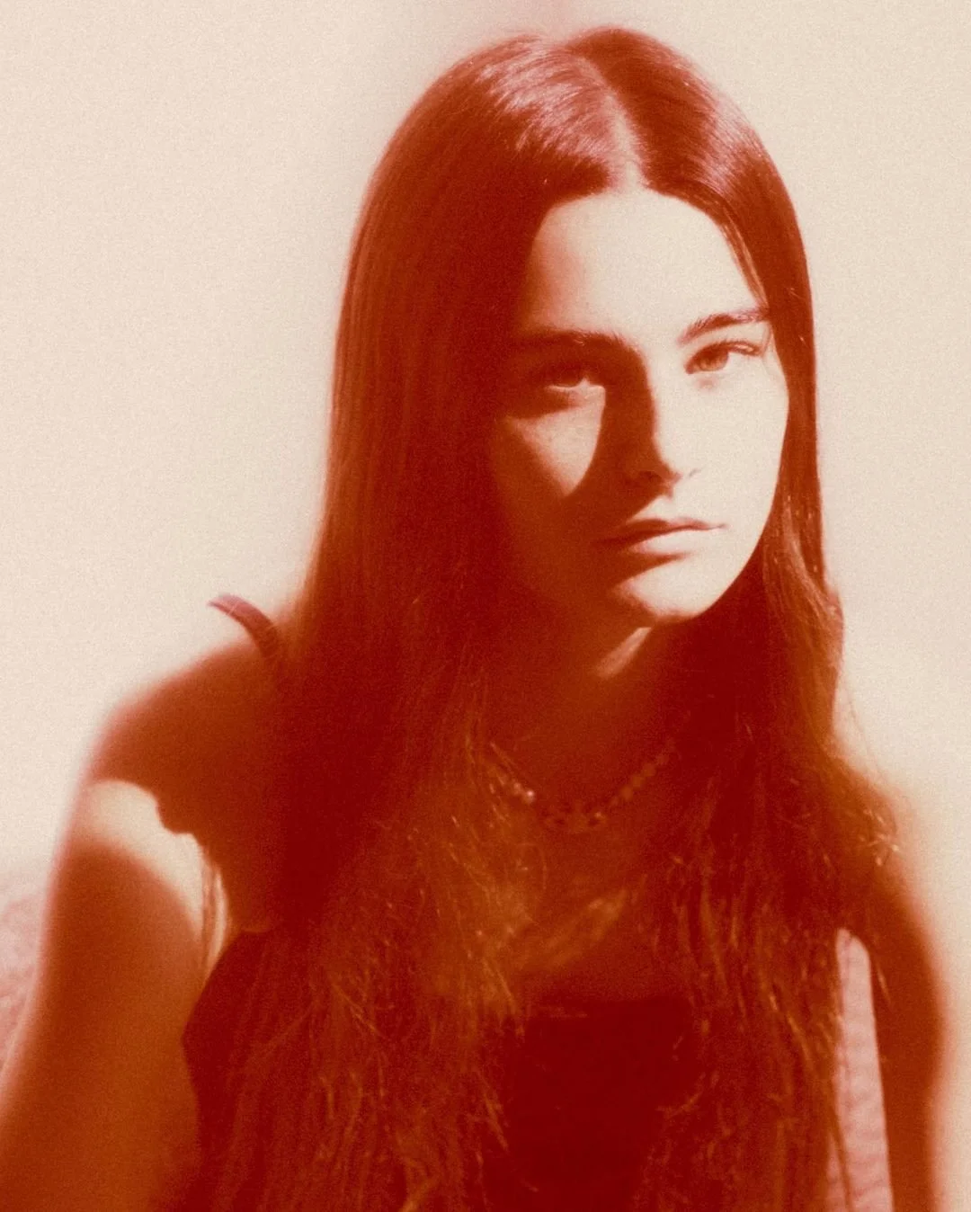 A woman with long red hair, wearing a necklace, looking at the camera with a neutral expression, in monochrome reddish tint.