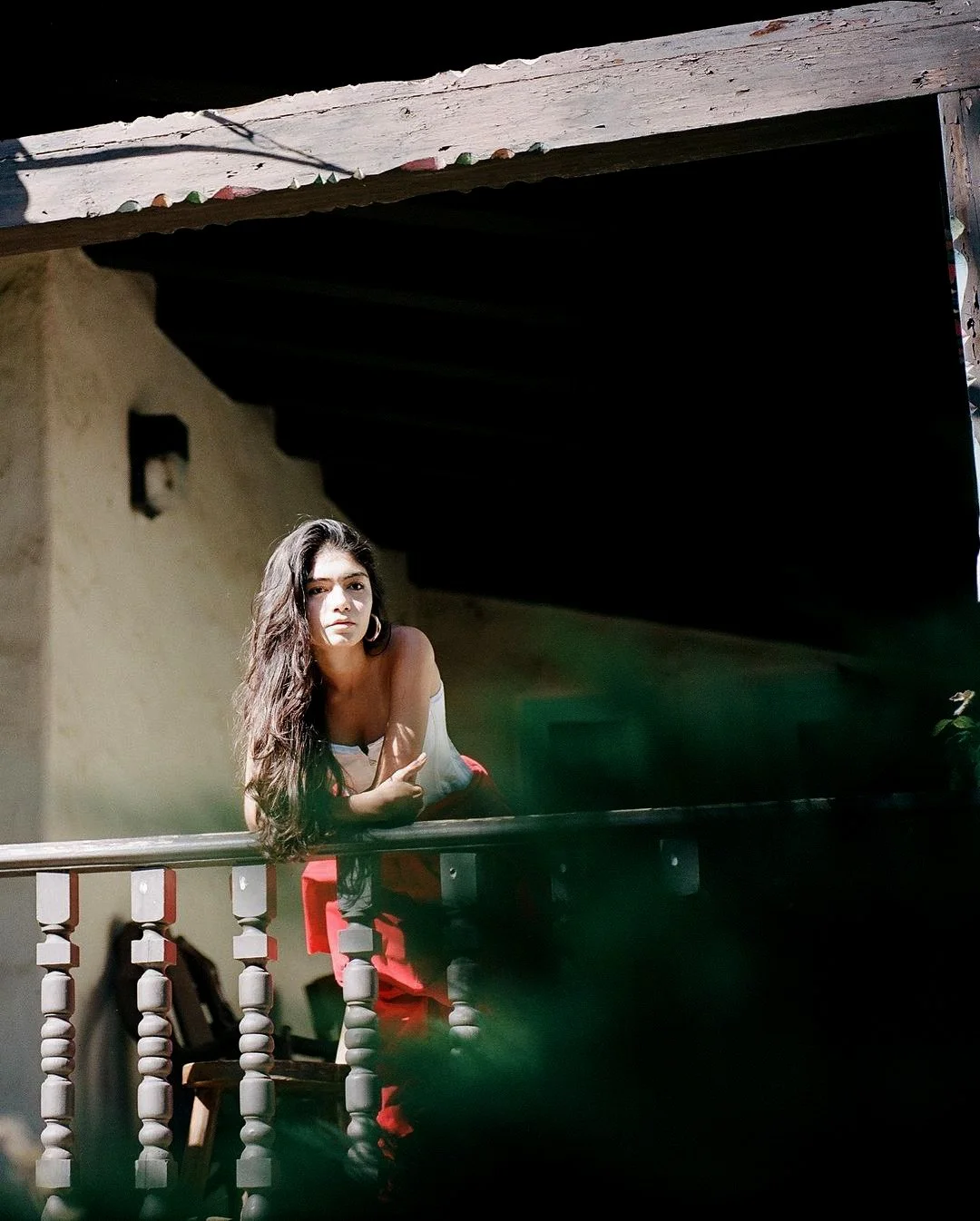 A woman with long dark hair leaning on a wooden railing on a porch, looking into the camera with a neutral expression.