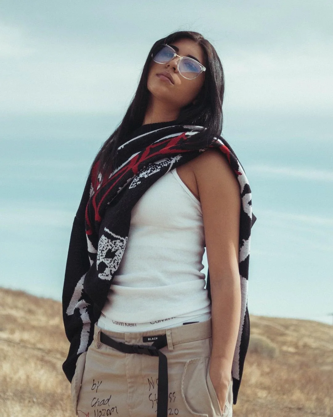 A woman with long dark hair wearing sunglasses, a white tank top, and beige pants standing outdoors on a rocky hill with a cloudy sky and ocean in the background, holding a black and red patterned jacket over her shoulder.