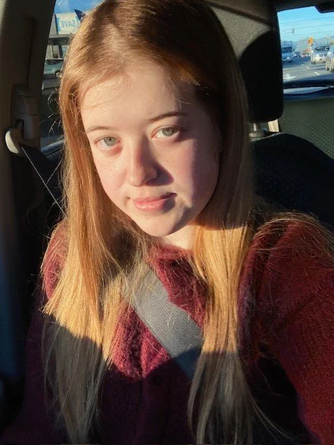 A young girl with long, light brown hair and blue eyes sitting in a car. She is wearing a maroon sweater and a seatbelt.