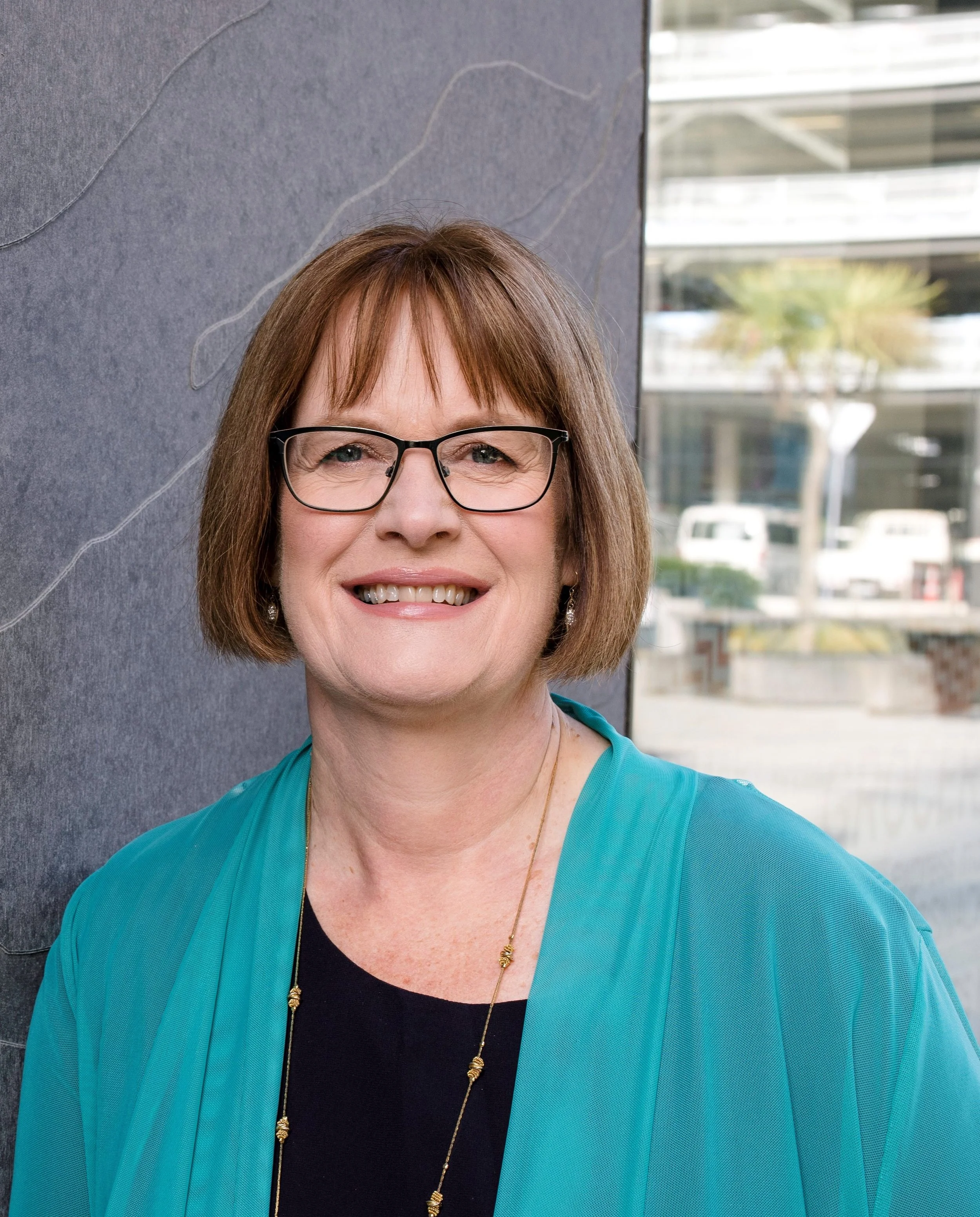 woman with short brown hair, wearing glasses, a turquoise jacket, a black top, and a gold necklace, standing outdoors against a gray wall with a cityscape background.